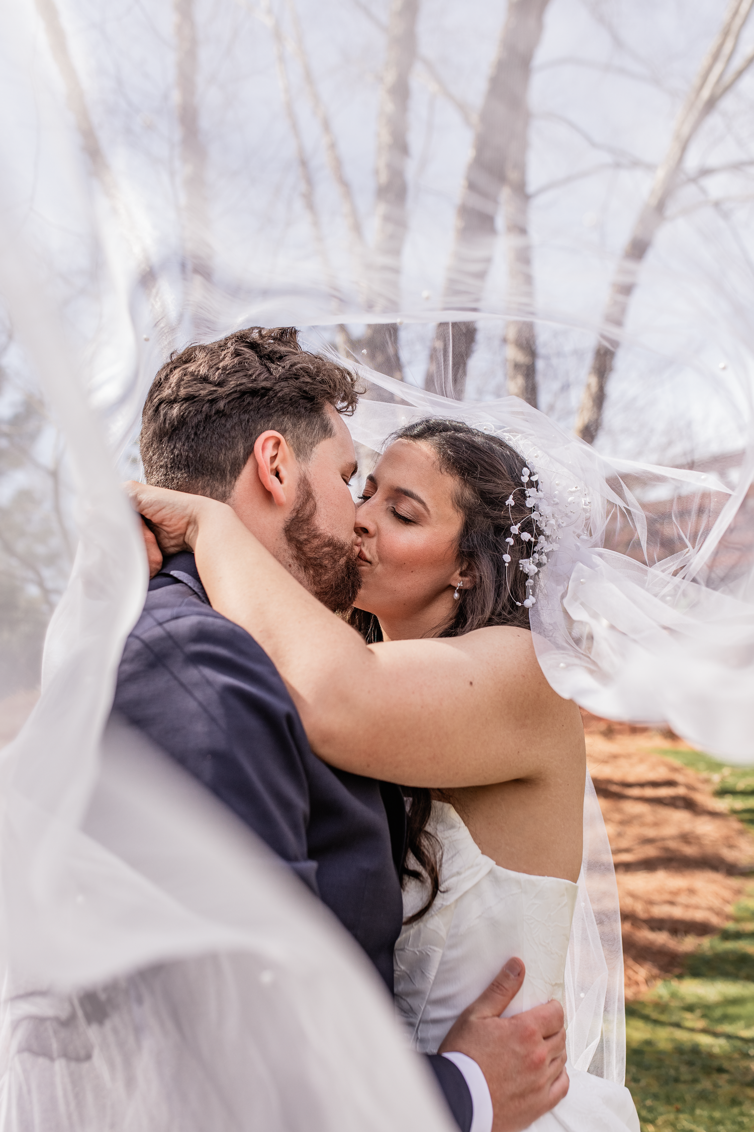 Bride and groom share a romantic kiss under a flowing veil in a misty outdoor winter wedding setting.