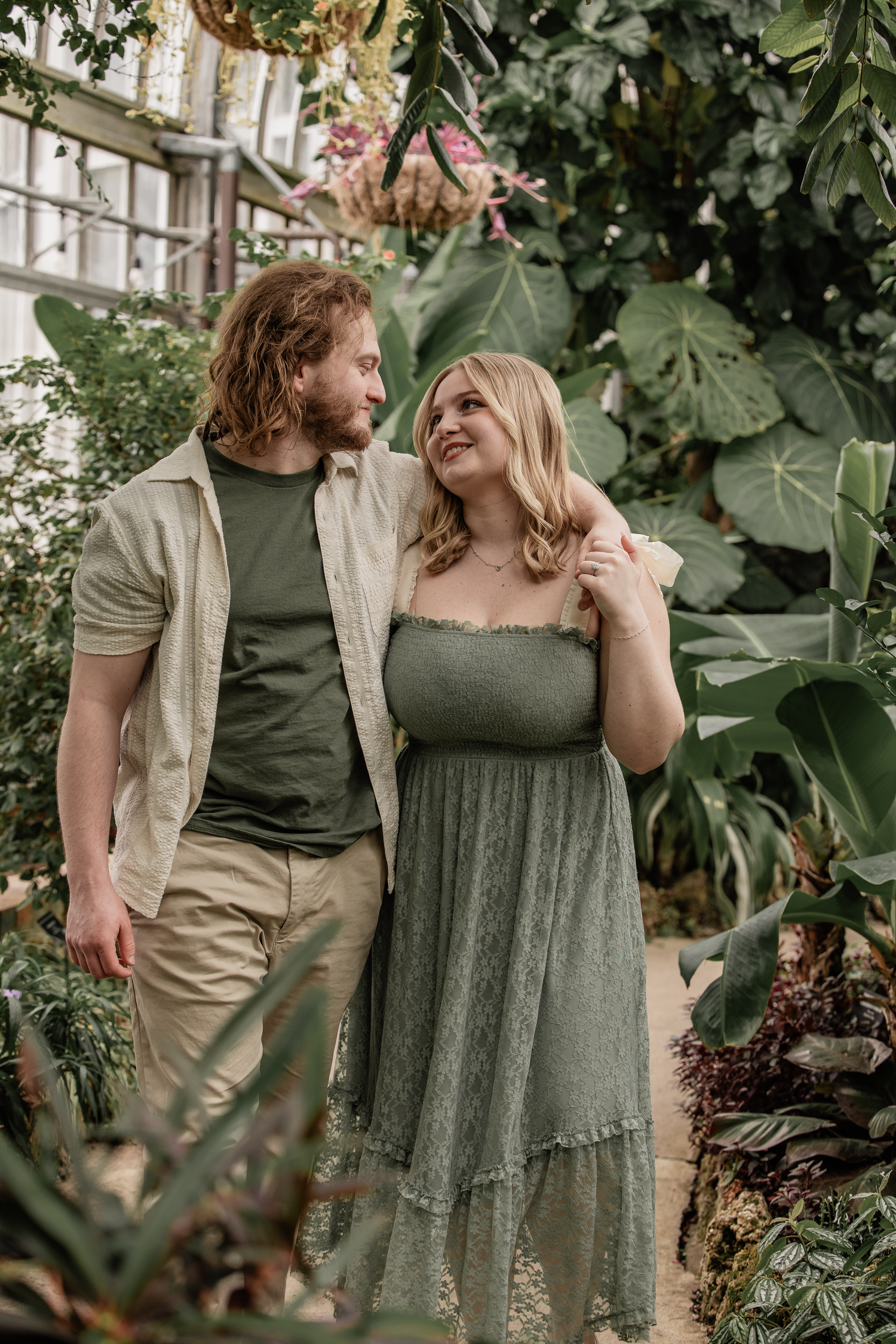 A couple walks arm in arm through a lush greenhouse, wearing coordinating green outfits, smiling at each other.