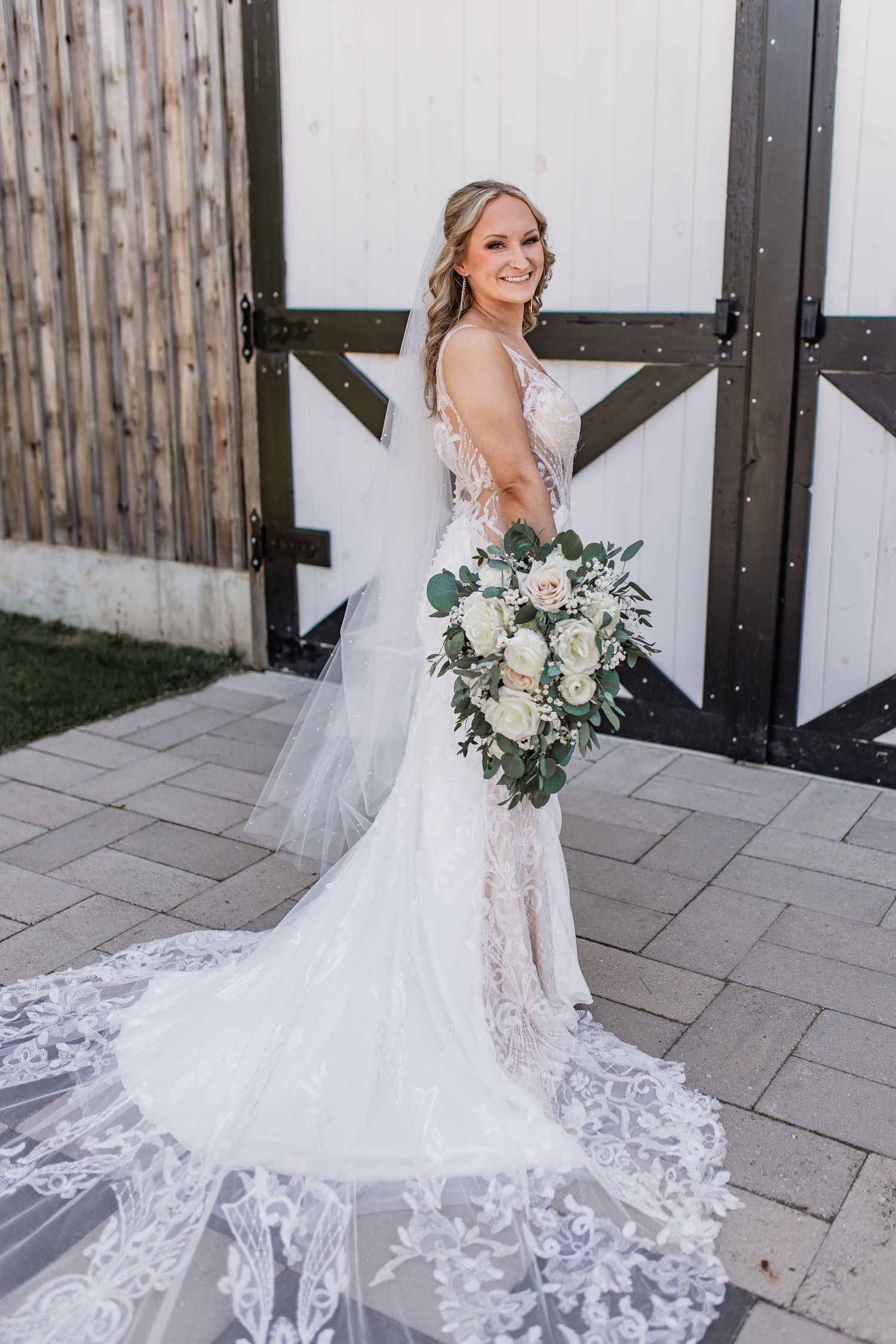 A bride in a stunning lace wedding gown with a flowing train poses elegantly on stone pavers outside a barn, for mid michigan local wedding
