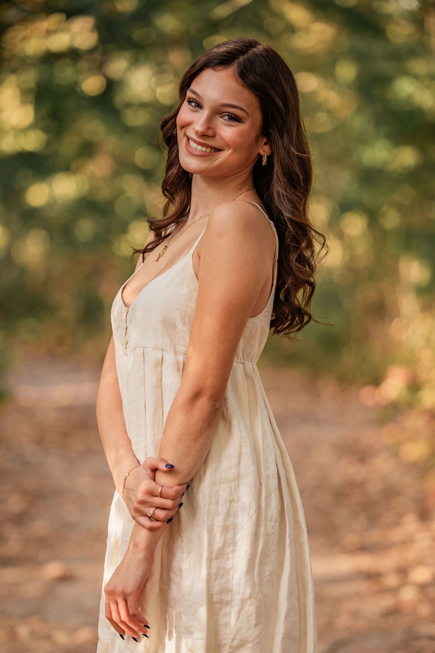Young woman in white sundress smiling outdoors in a sunlit forest path with bokeh green background.