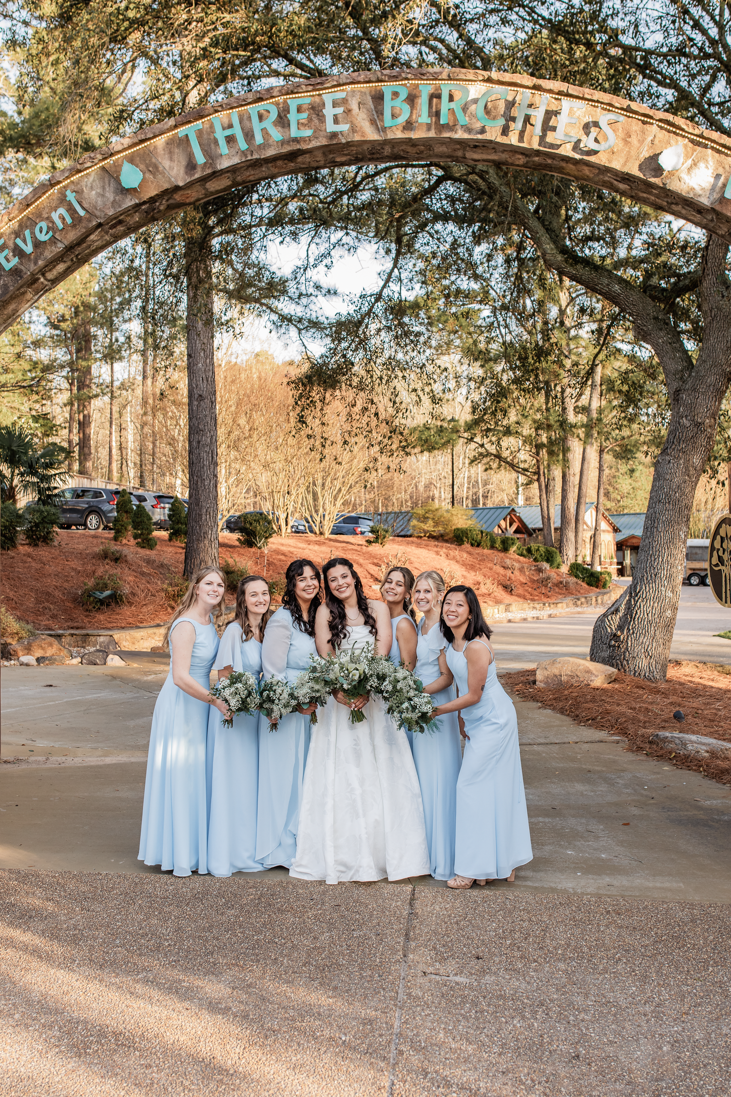 Bride and bridesmaids in light blue dresses pose under Three Birches Event venue arch, holding bouquets outdoors, wedding photographer