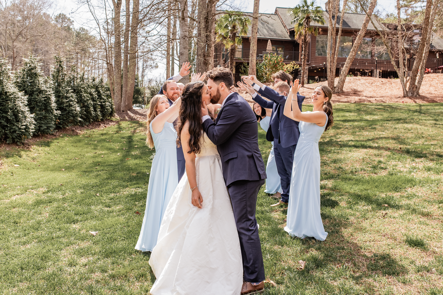 Bride and groom share first kiss surrounded by bridesmaids in light blue dresses and groomsmen outdoors, local michigan wedding photographer