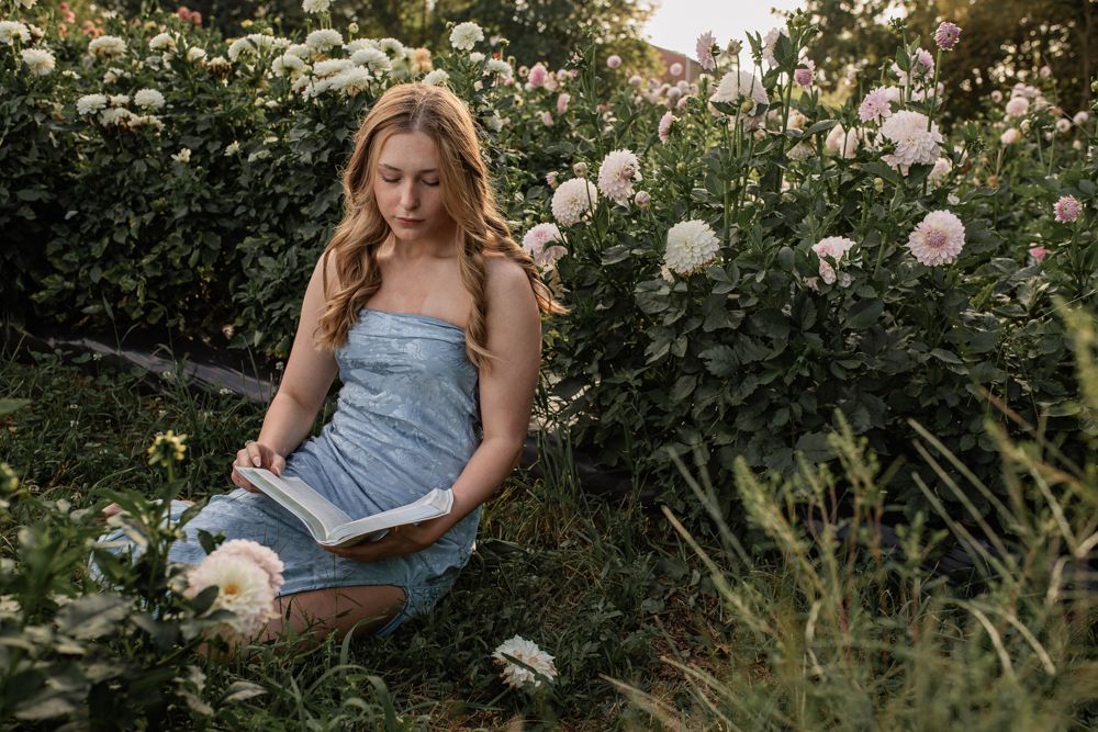 Young blonde woman in blue dress reading a book while sitting in a lush garden with blooming white and pink flowers.