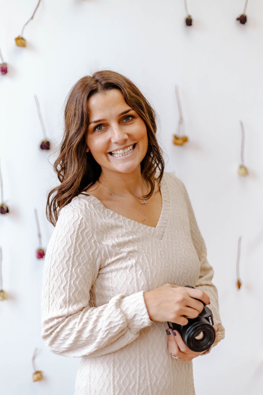 Professional photographer in cream sweater holds camera against white backdrop with hanging floral decorations.