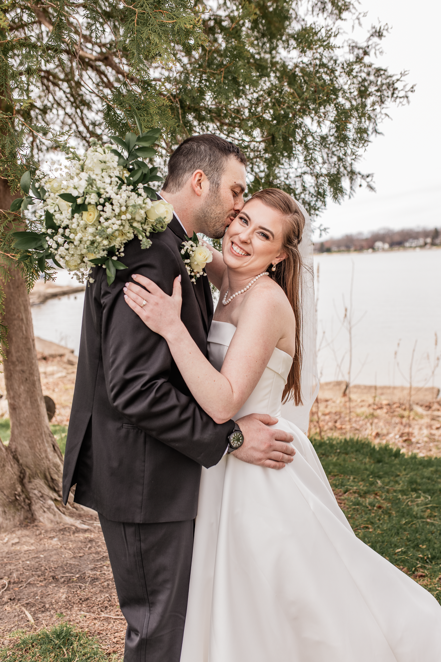 A bride and groom share a romantic embrace near a waterfront tree, grand rapids michigan photographer, mid michigan wedding photographer