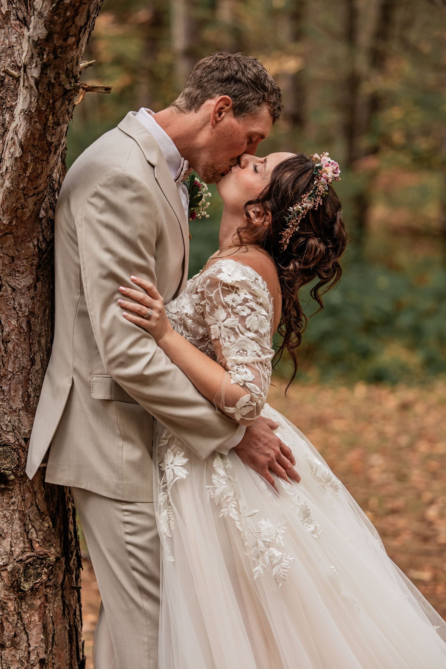 A bride and groom share a romantic kiss against a tree in an autumn forest, wearing elegant wedding attire.