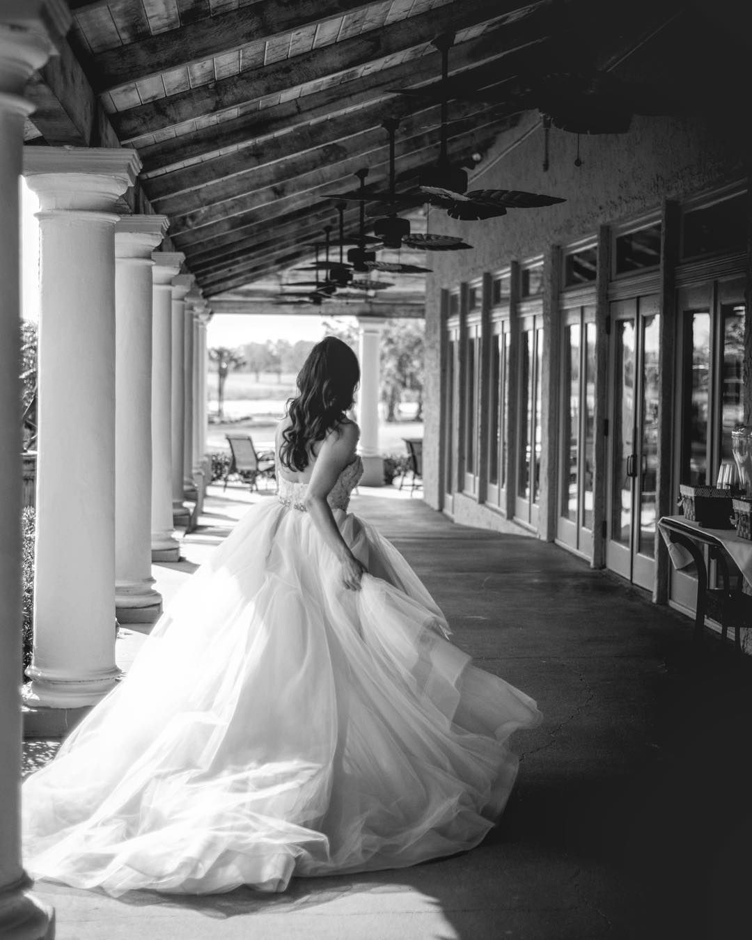 Black and white photo of a bride in a flowing tulle gown walking along a columned covered porch with ceiling fans.