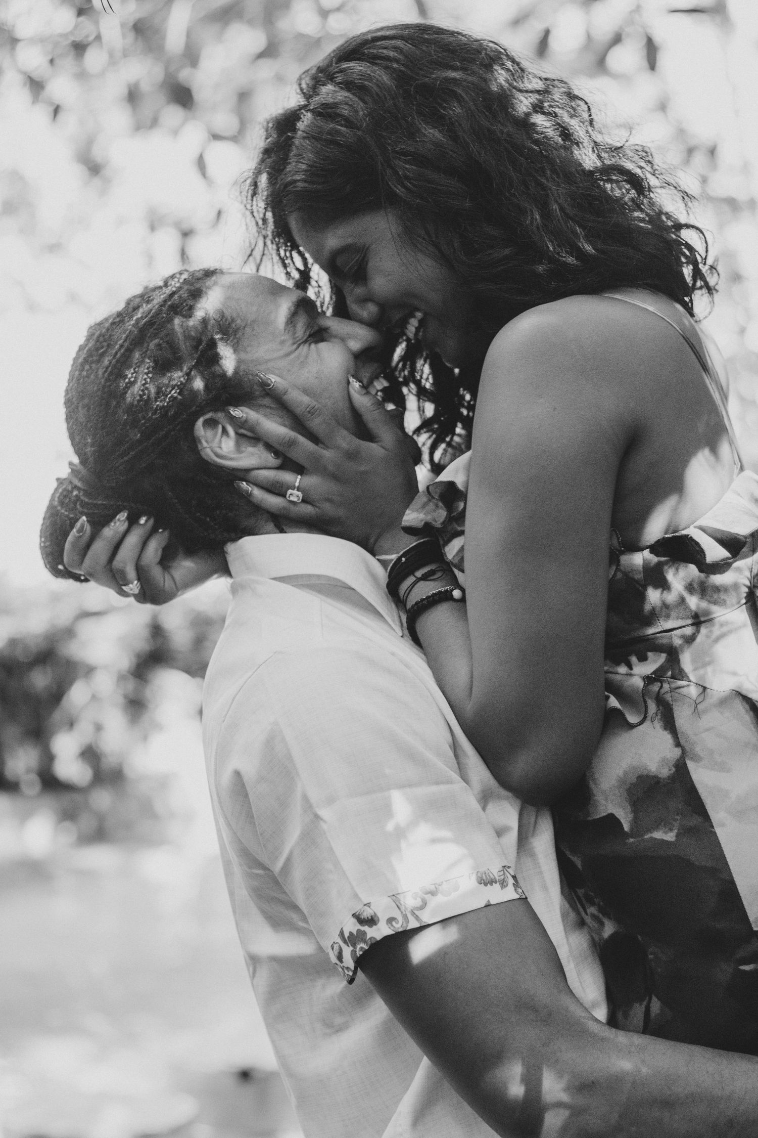 Black and white photo of a couple embracing outdoors as a woman in a floral dress is lifted and kisses a smiling man.