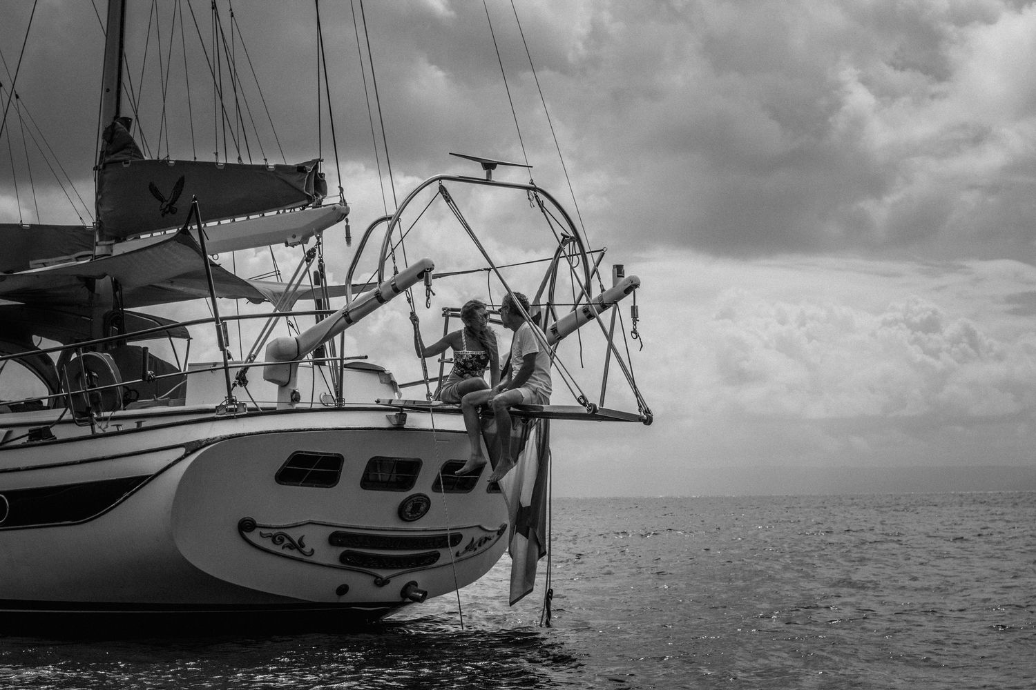 Black and white photo of two people sitting at the bow of a sailboat on calm water beneath a dramatic cloudy sky.