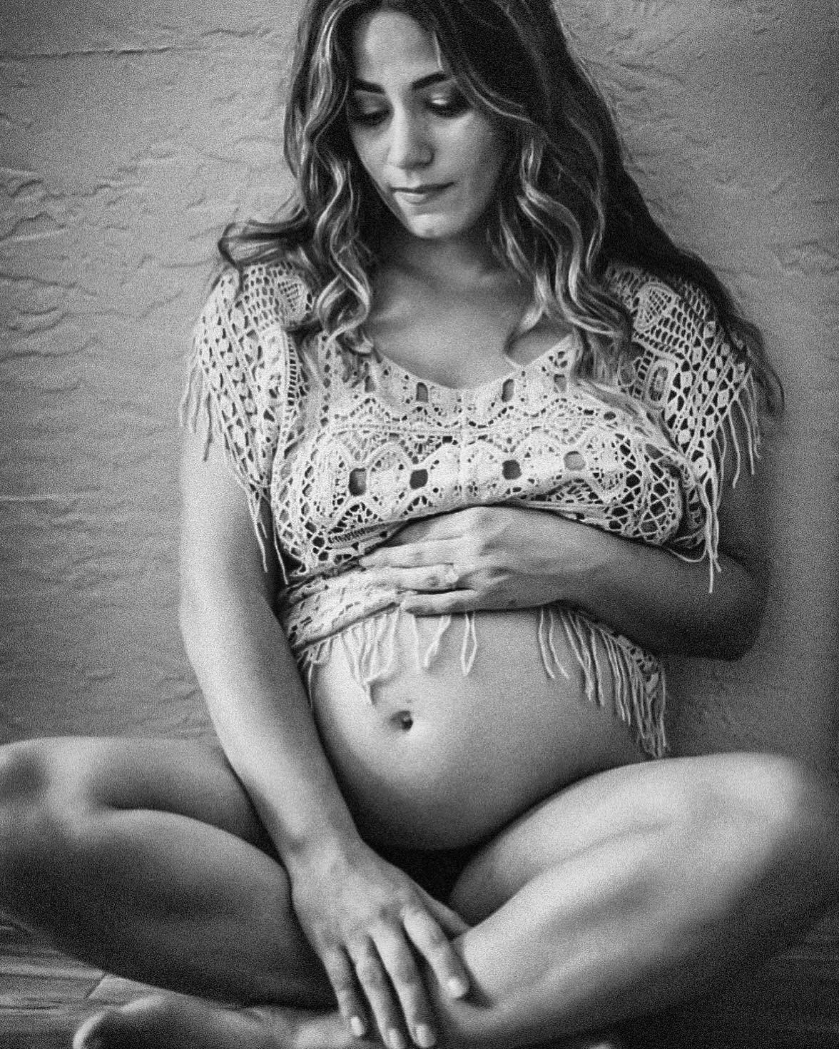 Black and white maternity portrait of an expectant mother in a crochet top sitting cross-legged and cradling her baby bump.