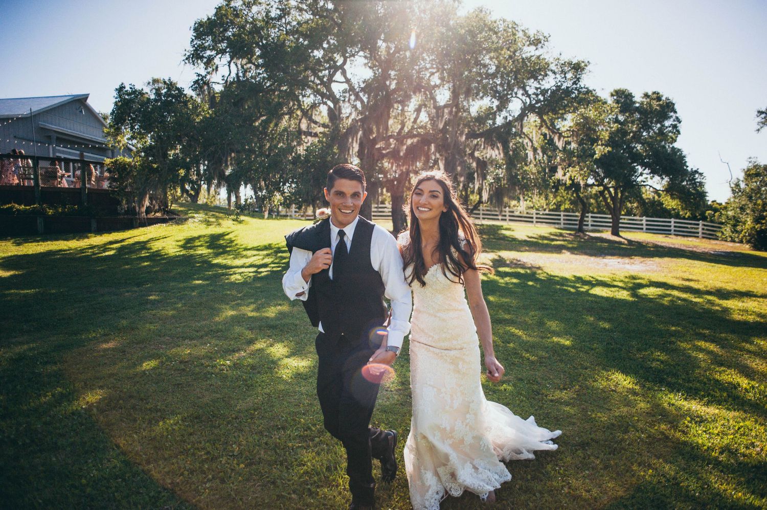 Bride in a lace wedding gown and groom in a vest hold hands and walk across a sunlit lawn beneath large oak trees.
