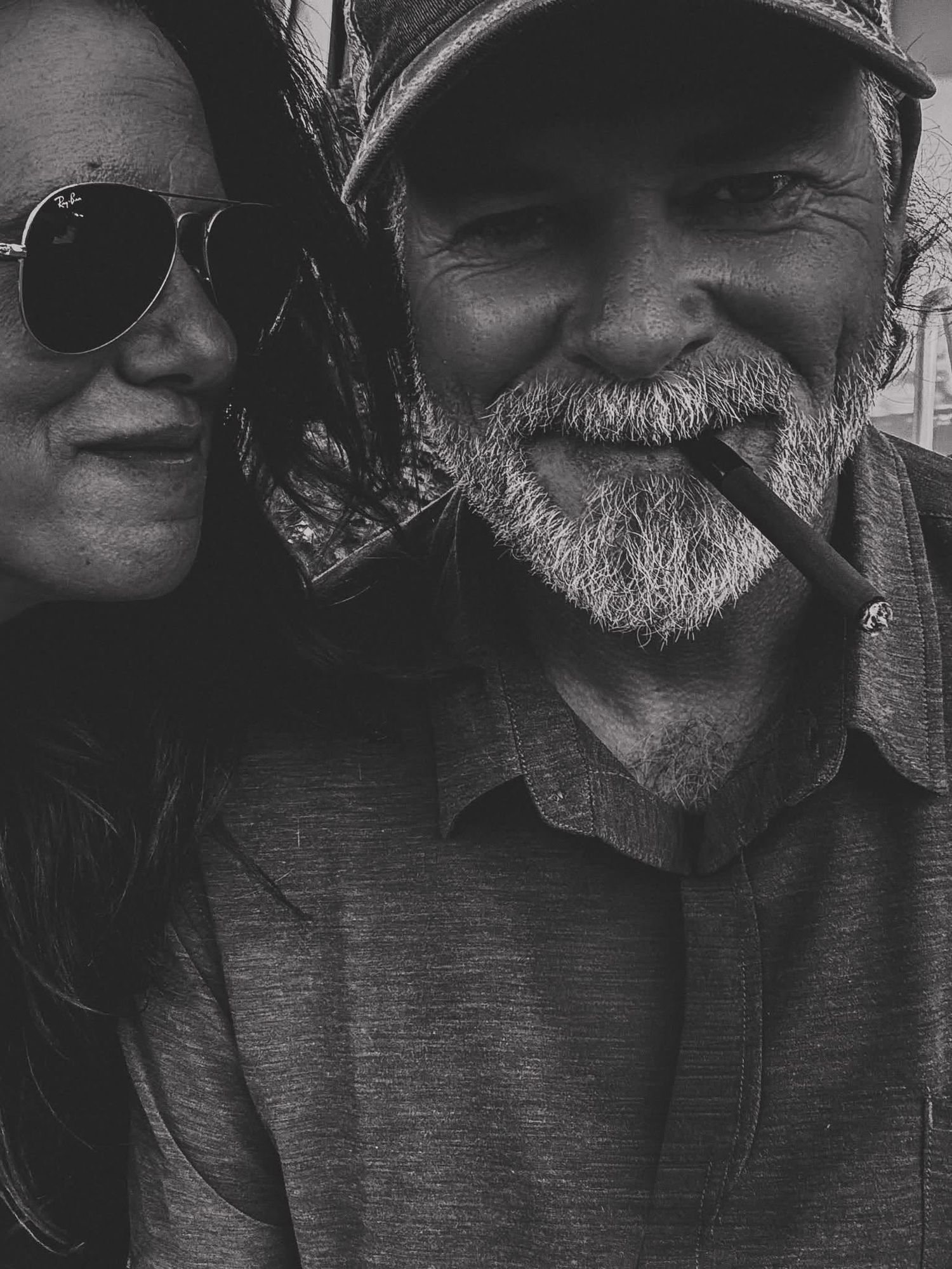 Black and white selfie of a smiling bearded man with a cigar and a woman in aviator sunglasses leaning in close.