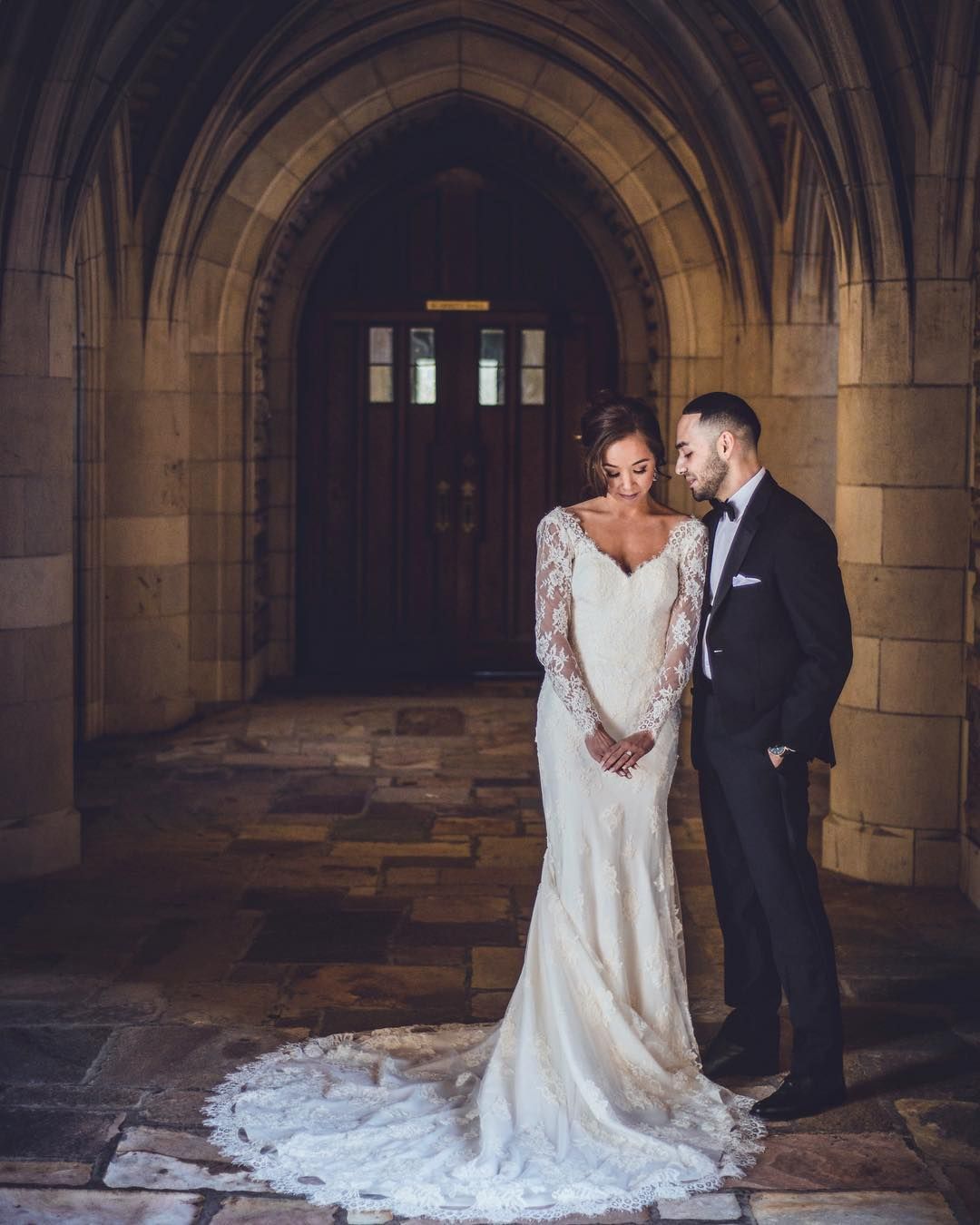 A bride in a lace gown and a groom in a dark suit stand together in a stone archway with wooden doors.