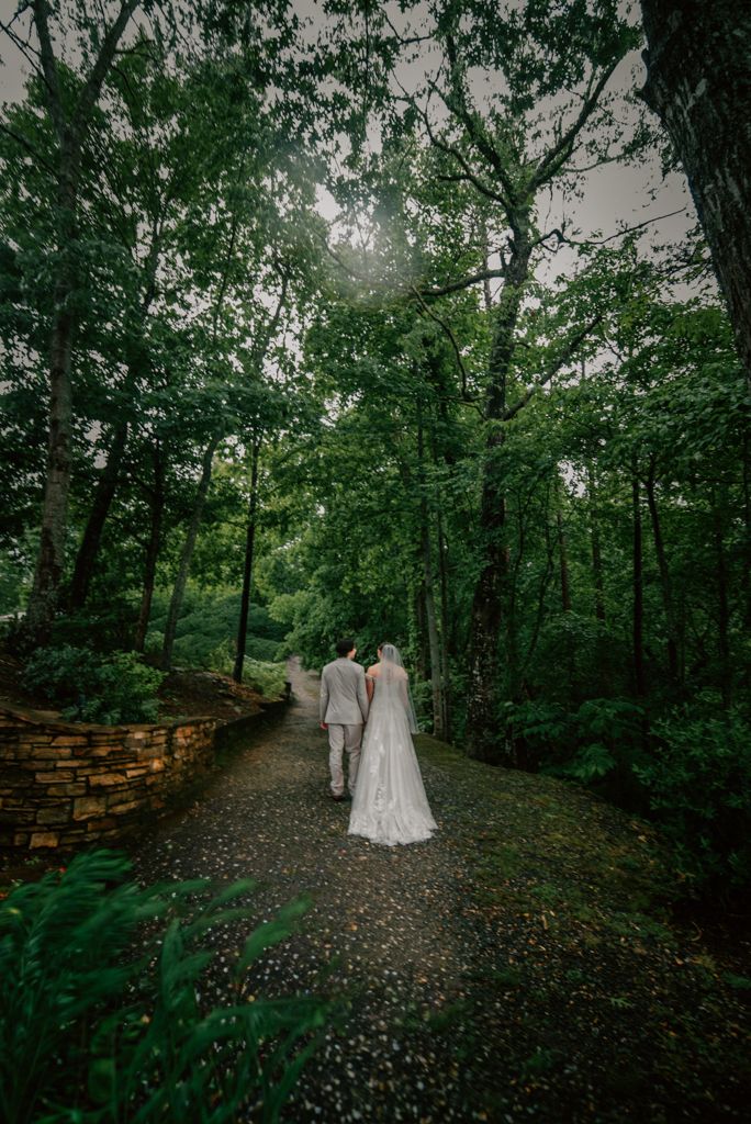 Bride in a long white gown and groom in a light suit walk a gravel path through a lush green forest on their wedding day.