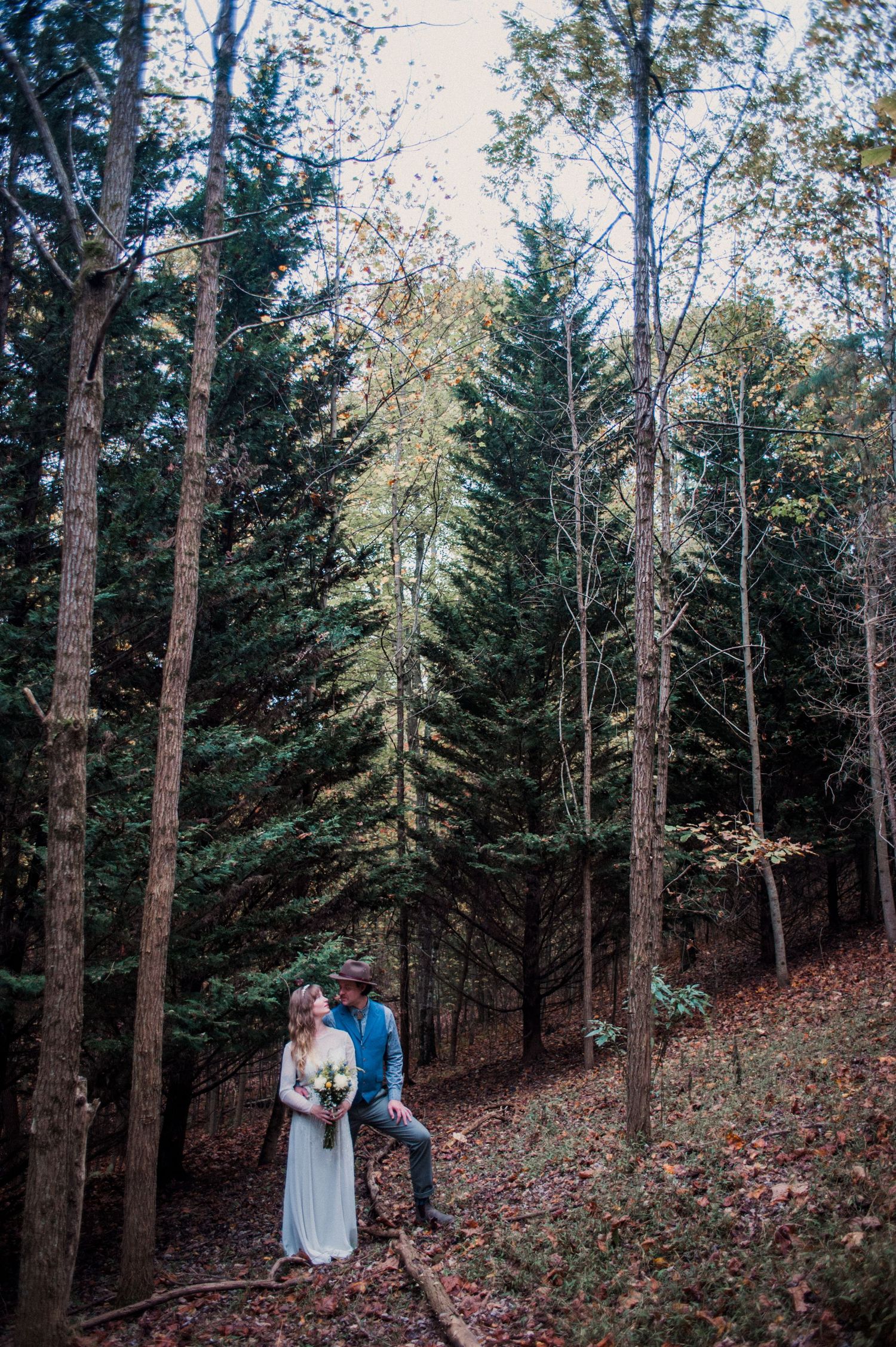 A bride in a white dress holding a bouquet and a groom in a hat stand among tall pine trees on a leafy forest hillside.