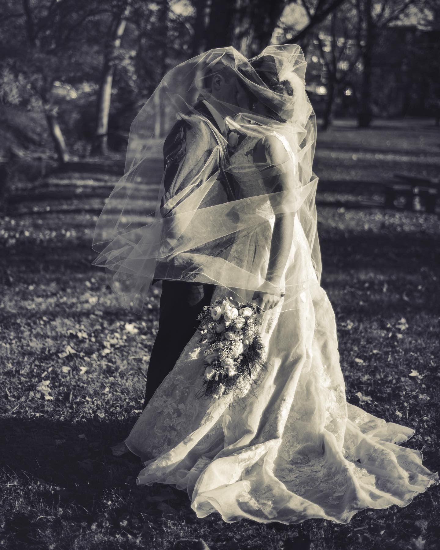 Black and white portrait of a bride and groom kissing beneath a sheer wedding veil, with the bride holding a floral bouquet.