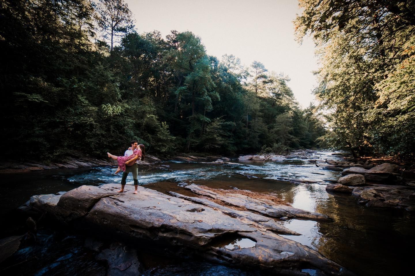 Groom lifting his bride in a joyful spin on a large flat rock in a river surrounded by lush green trees at golden hour.