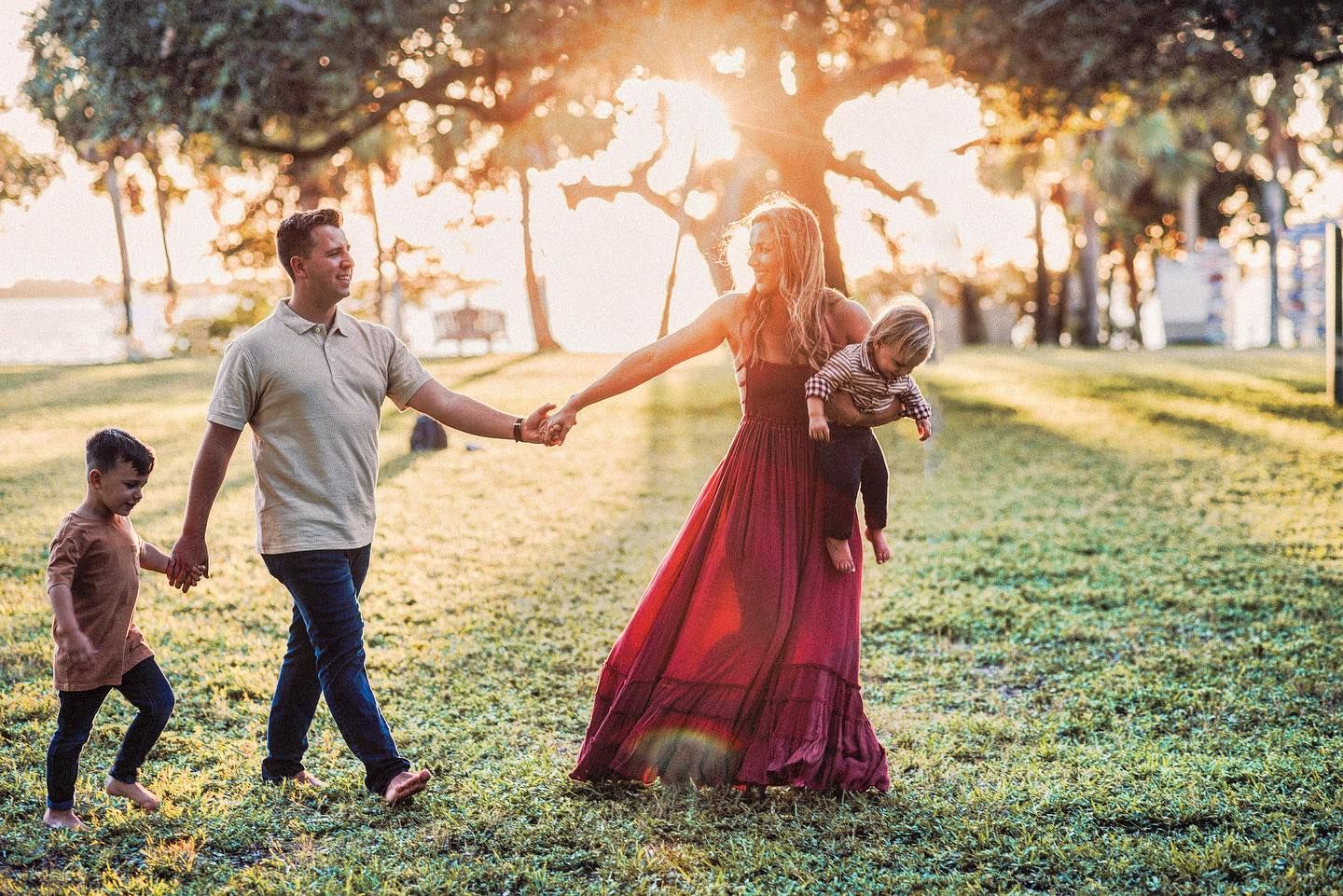Family of four holding hands and walking across a grassy park at sunset, with a mother in a red dress carrying a baby.