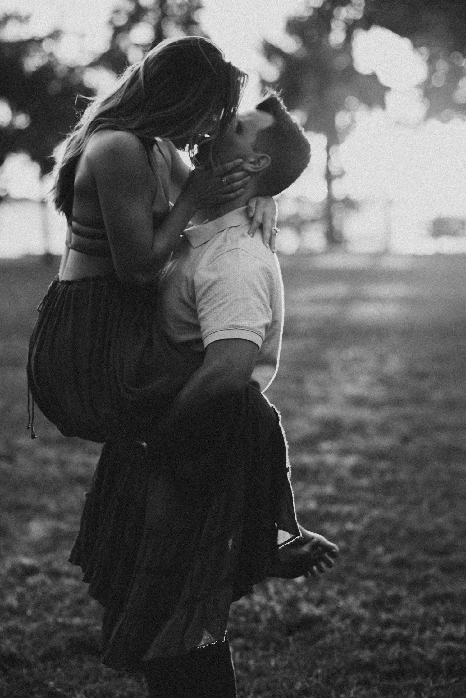 A black and white portrait of a man lifting a woman in a long skirt as they kiss in a softly lit park.