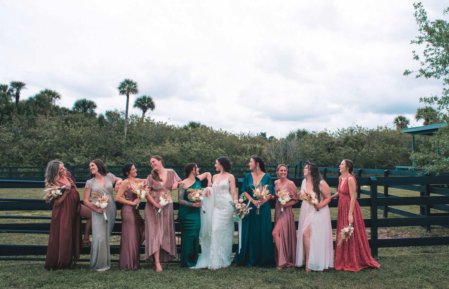 Bridesmaids in mismatched mauve, teal and blush dresses stand laughing beside the bride in white along a wooden fence.