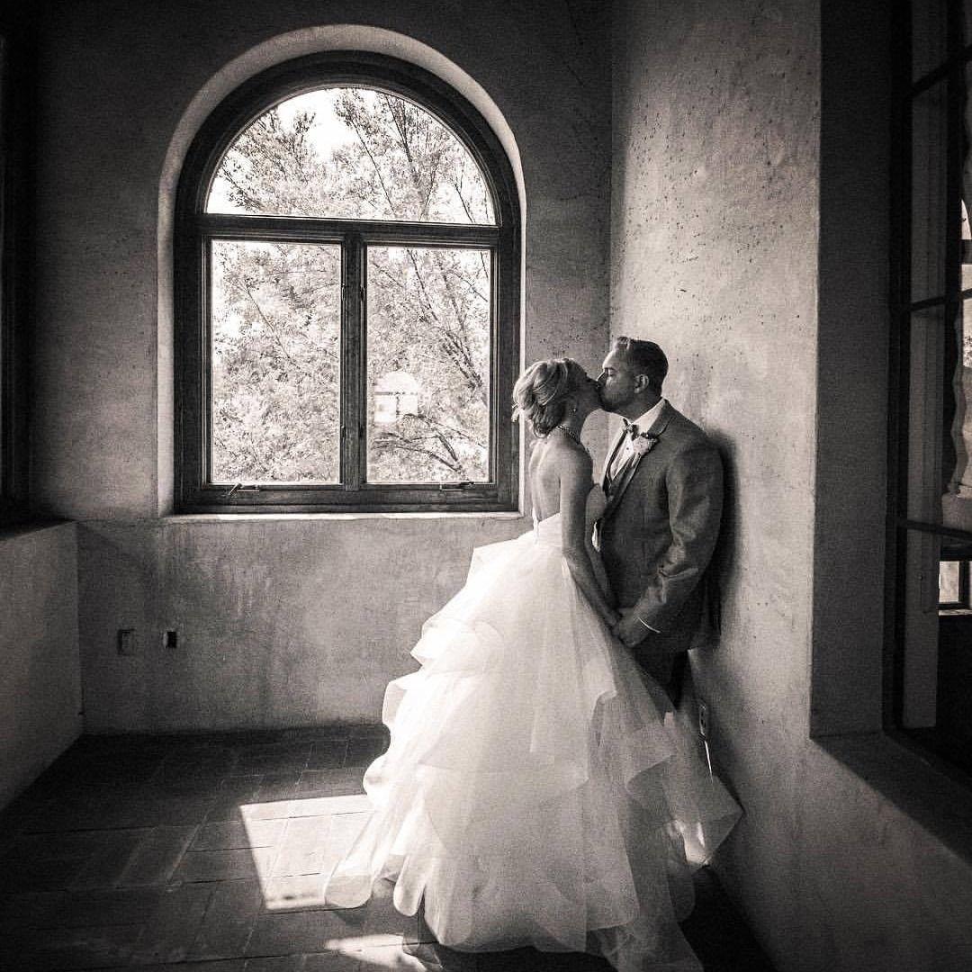 Groom kissing his bride's forehead beside a large arched window in a rustic room, her ruffled tulle wedding gown glowing in the light.