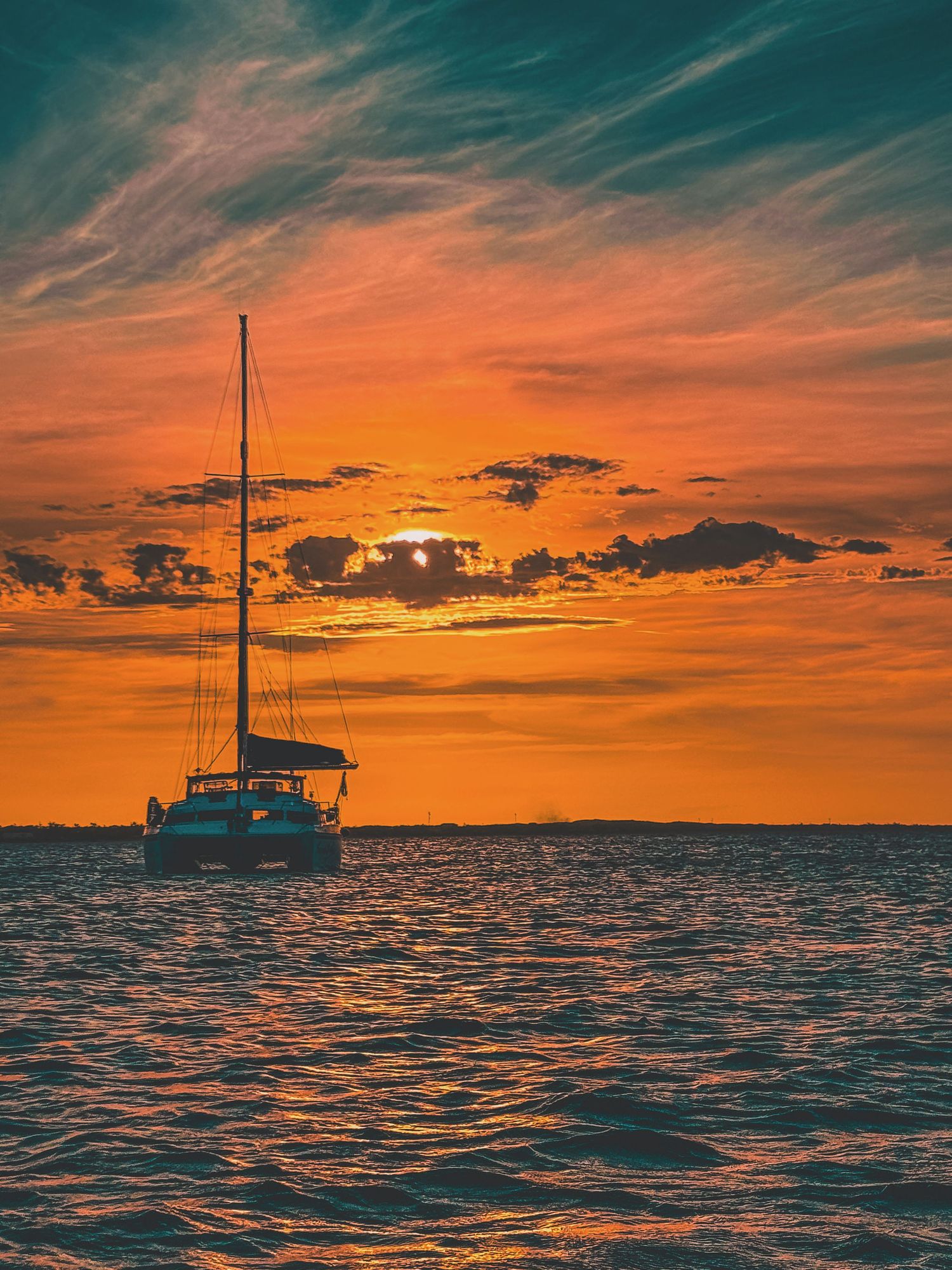 A sailboat anchored on rippling water silhouetted against a vivid orange and teal sunset sky.