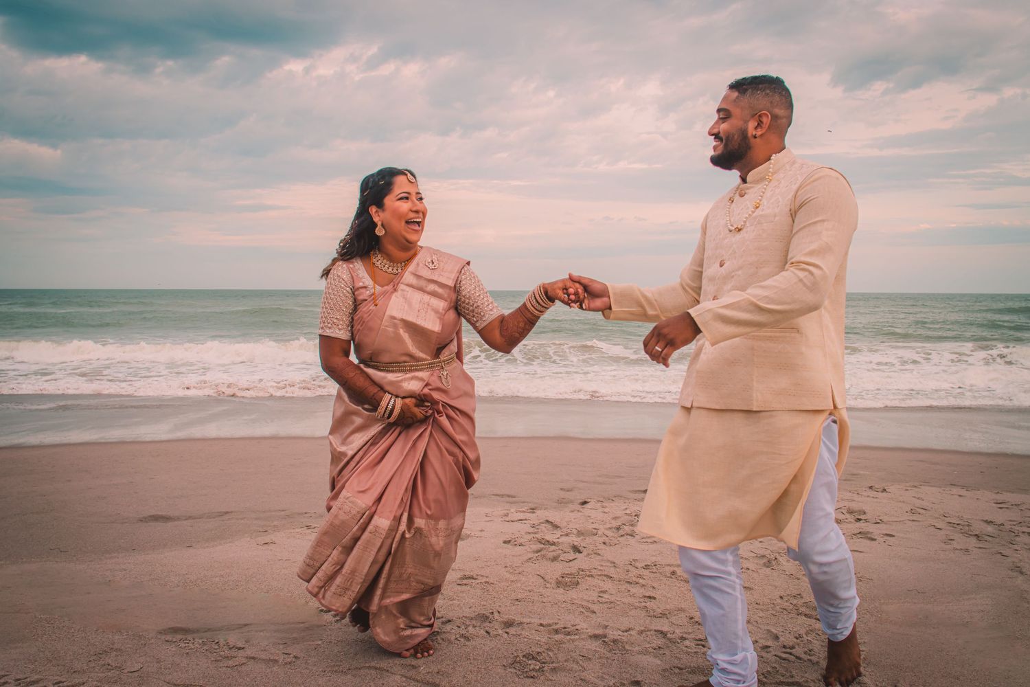 Smiling couple in traditional Indian attire hold hands and walk along a beach with ocean waves and a pastel sunset sky behind.