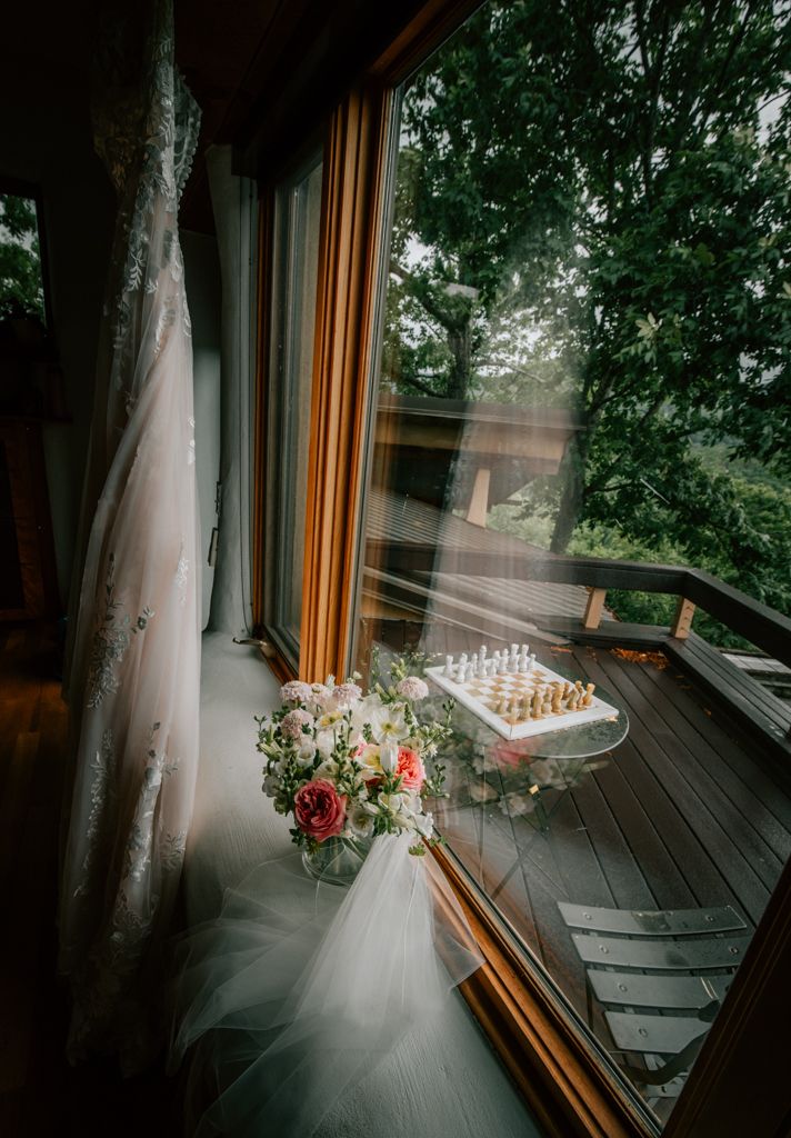 A bridal bouquet of pink roses sits on a window sill with a wedding veil and a chess set on the wooden deck outside.