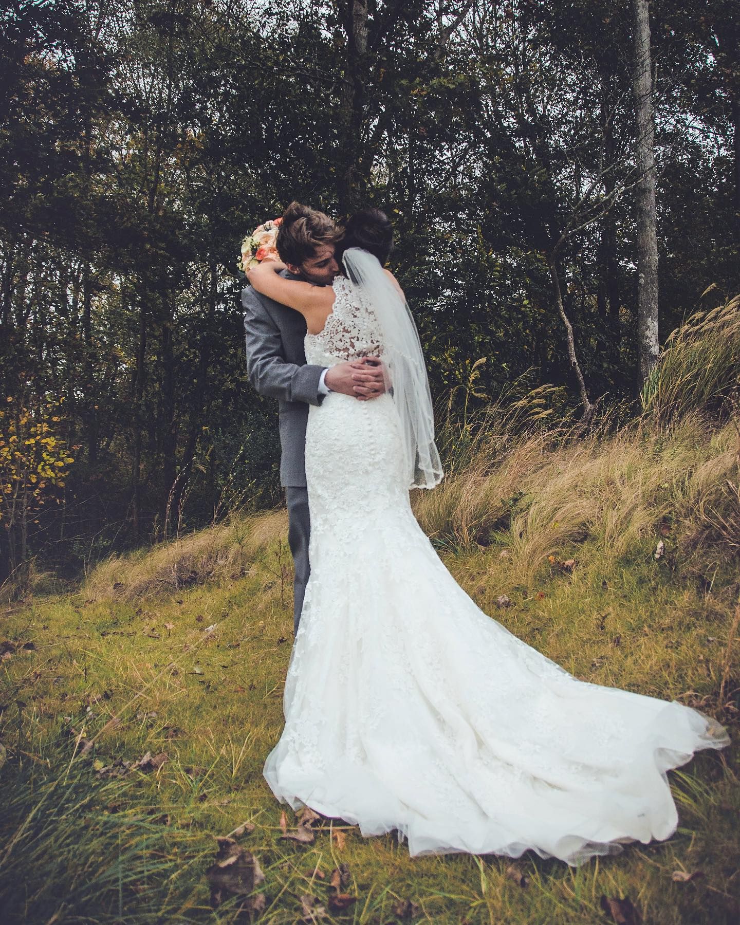 A bride in a white lace mermaid gown and veil embraces her groom on a grassy hillside surrounded by autumn woods.