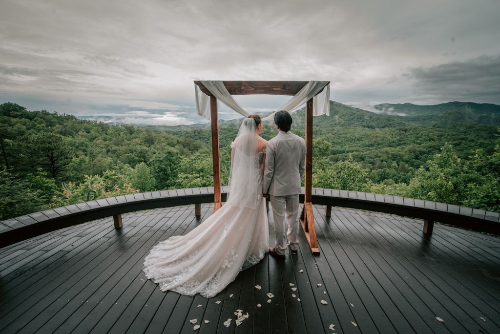A bride and groom stand under a wooden wedding canopy on a deck overlooking misty green mountains beneath a cloudy sky.