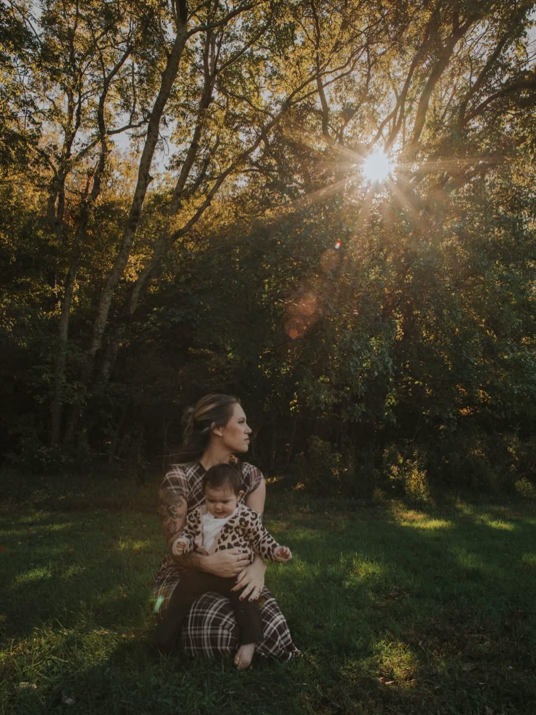 A mother in a plaid outfit holds her baby on a grassy lawn while golden sunlight streams through the autumn trees.