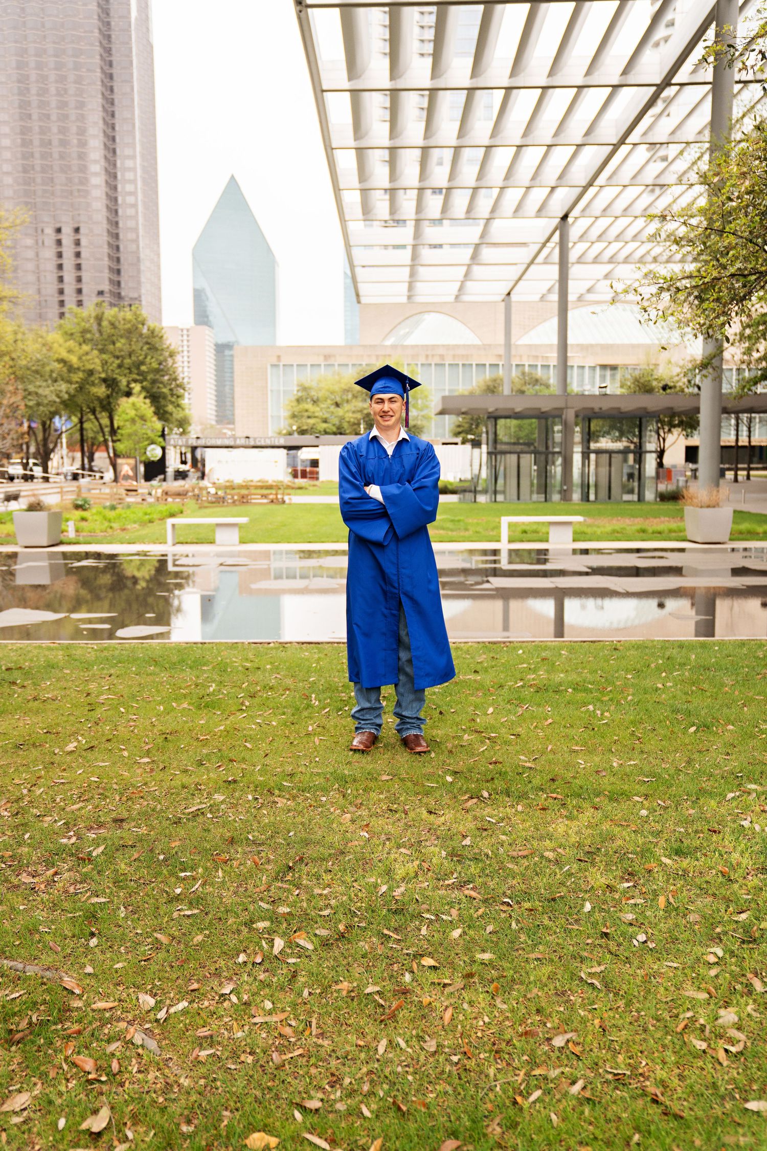 Graduate in blue cap and gown stands proudly with arms crossed on green lawn with city skyline backdrop.