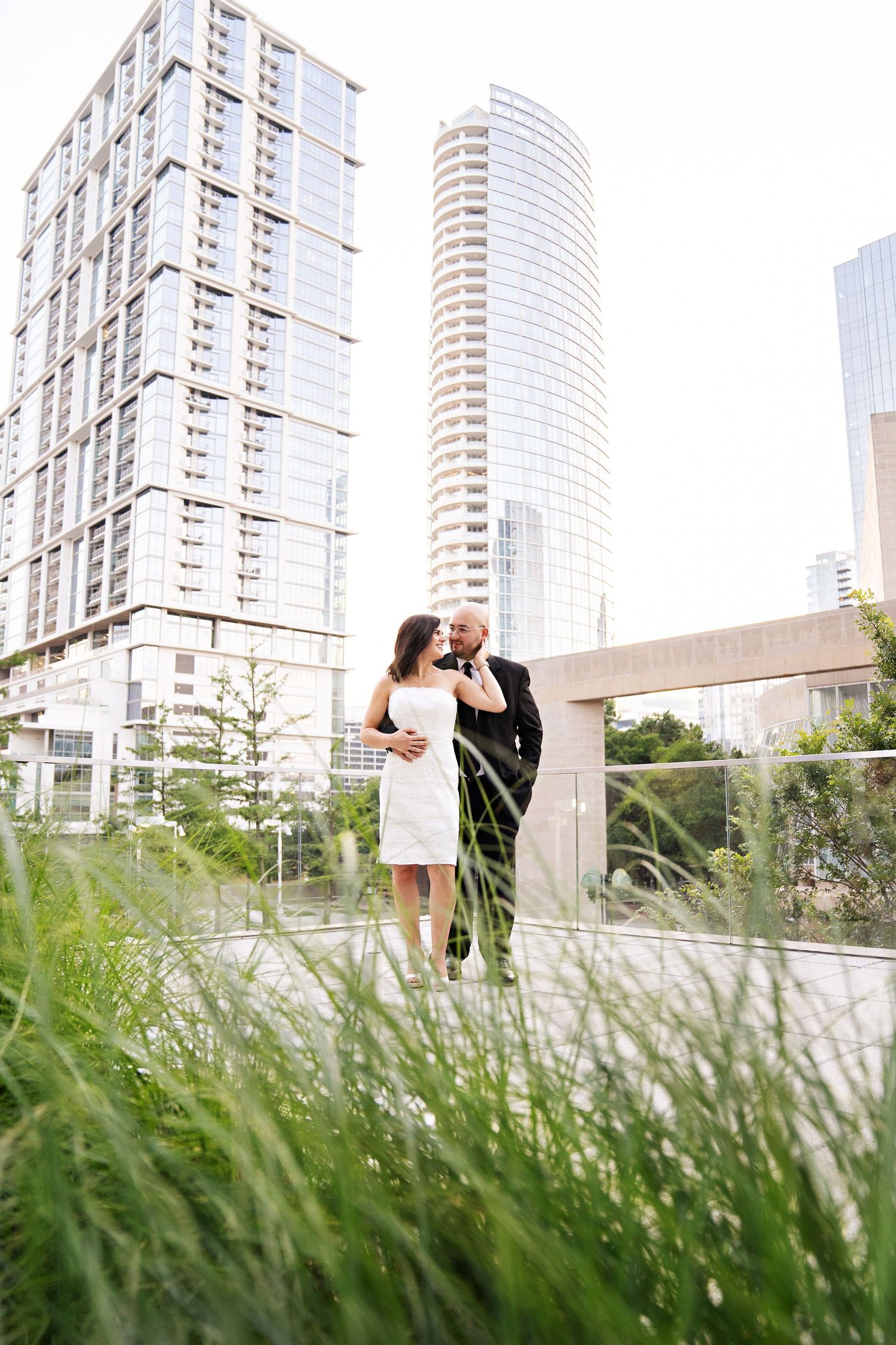 Couple embracing on urban rooftop garden with modern city skyscrapers in background, woman in white dress, man in dark suit.