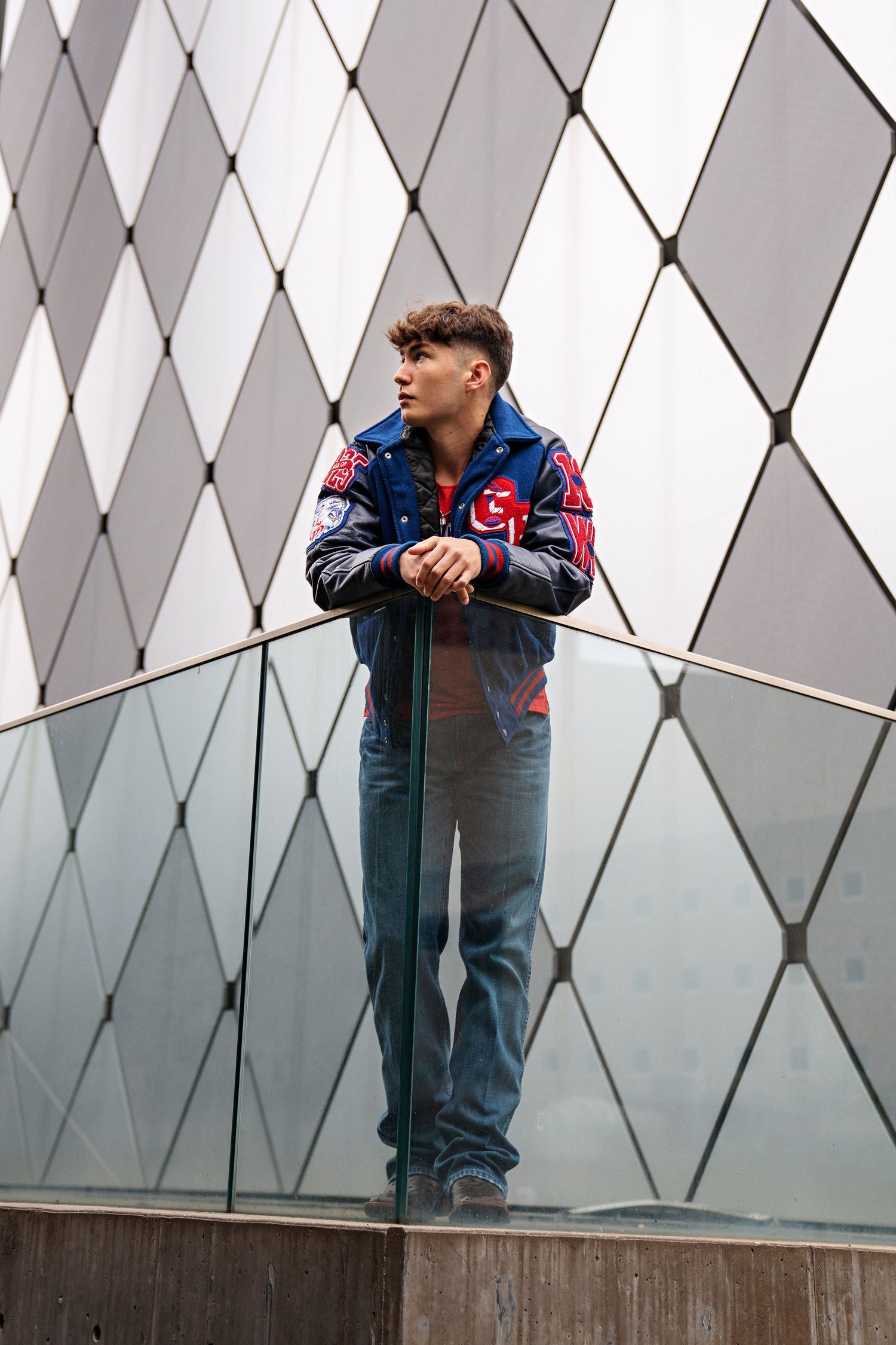 Young man in blue varsity jacket and jeans leaning on glass railing against geometric building facade.