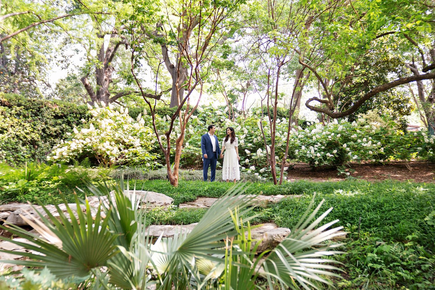 Couple standing hand in hand in a lush green garden with blooming white flowers and tropical plants.