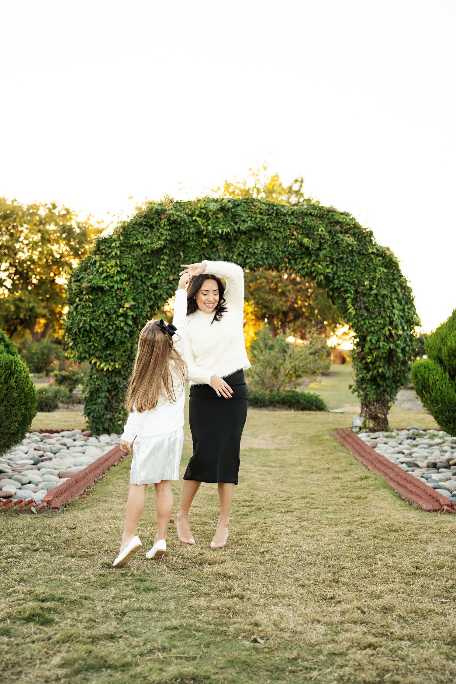 A mother and daughter dance together in a lush garden archway, both wearing white tops during golden hour.