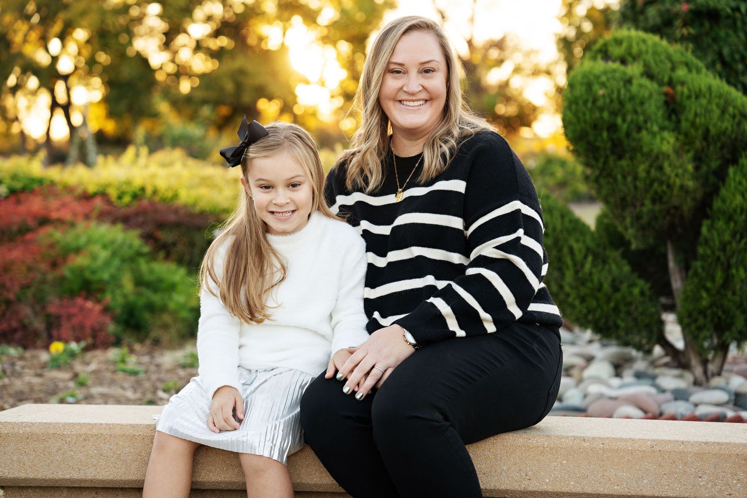 A woman and young girl in a white dress smile together on a stone bench in a park at golden hour.