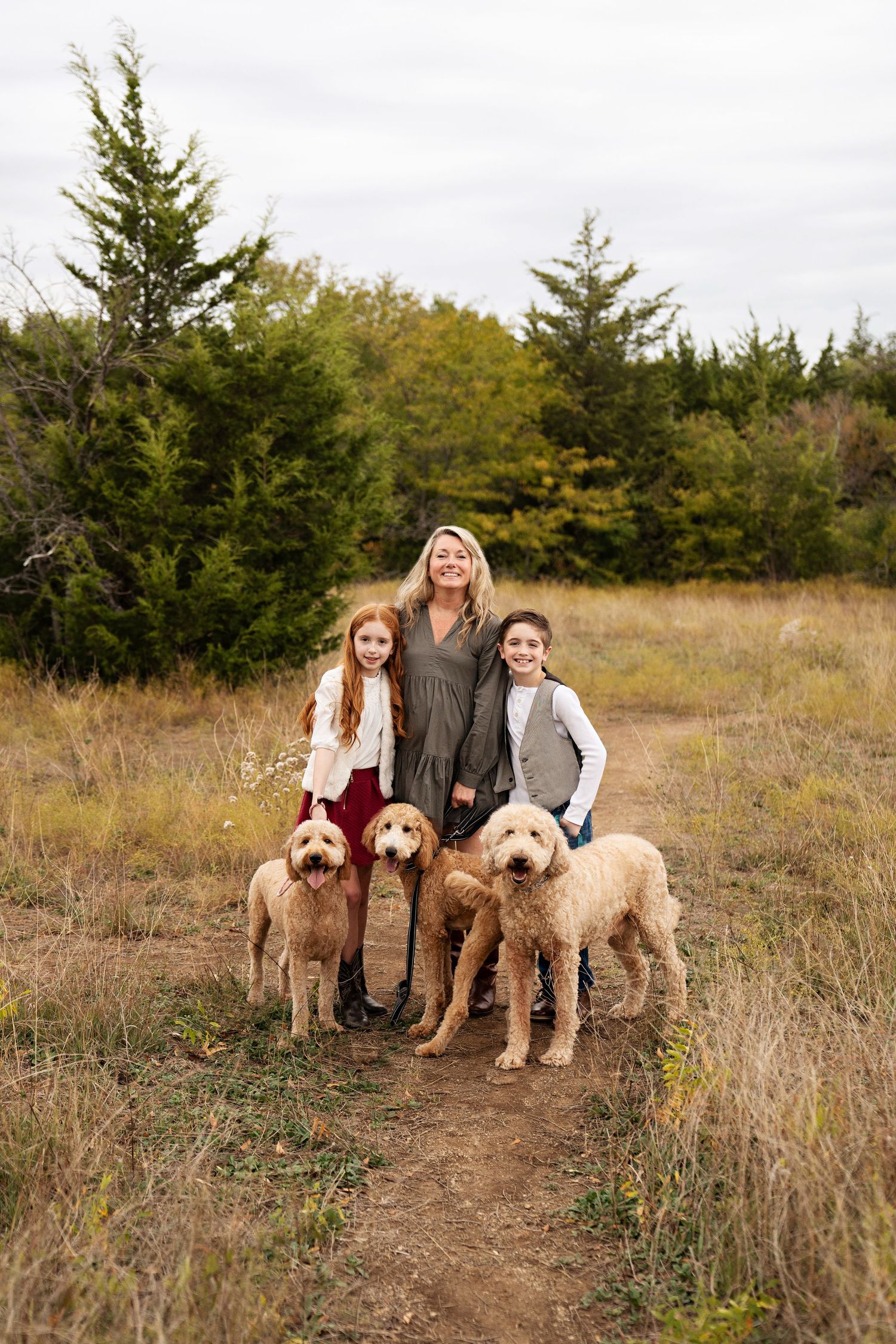 Woman and two children pose outdoors with three golden doodle dogs on a dirt path surrounded by tall grass and trees.