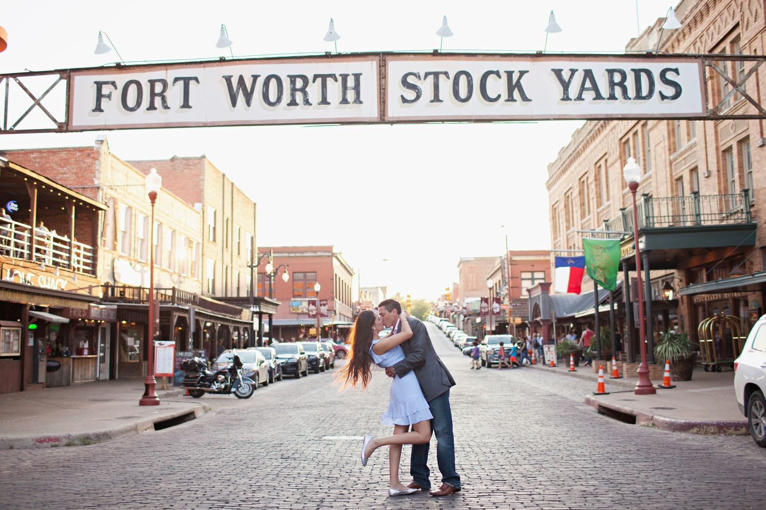 Couple kissing under Fort Worth Stock Yards sign on cobblestone street at dusk.