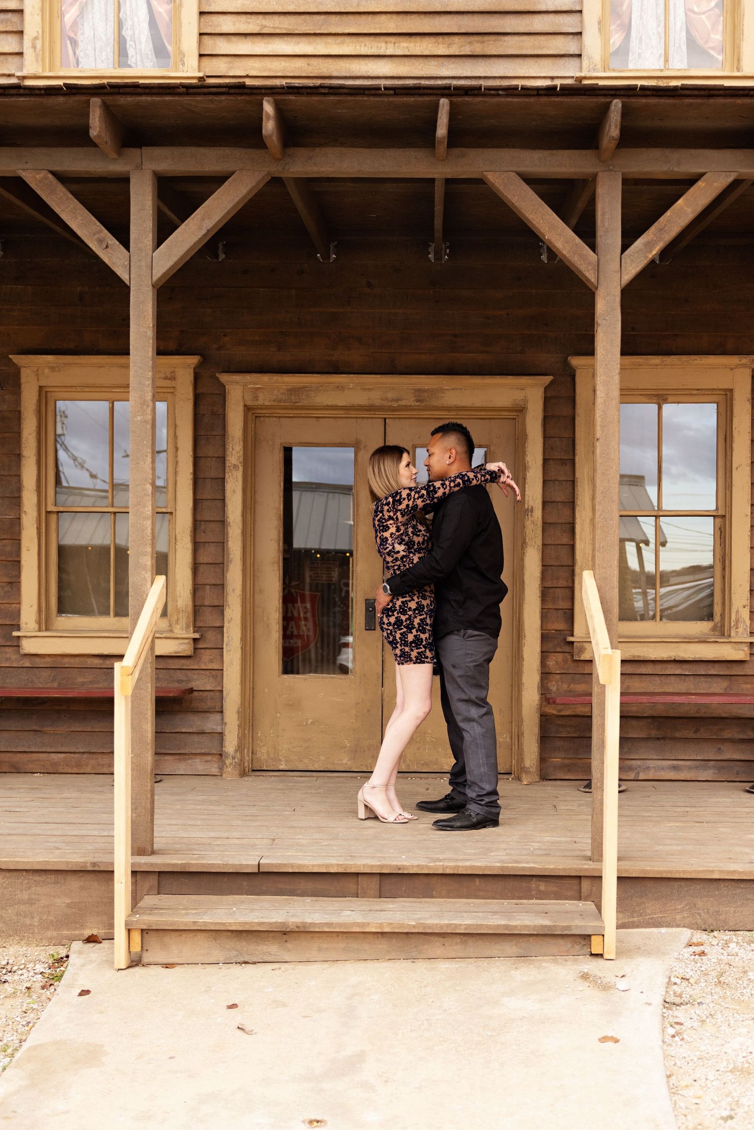 Couple embracing on wooden porch of rustic building, woman in floral dress and man in black suit.