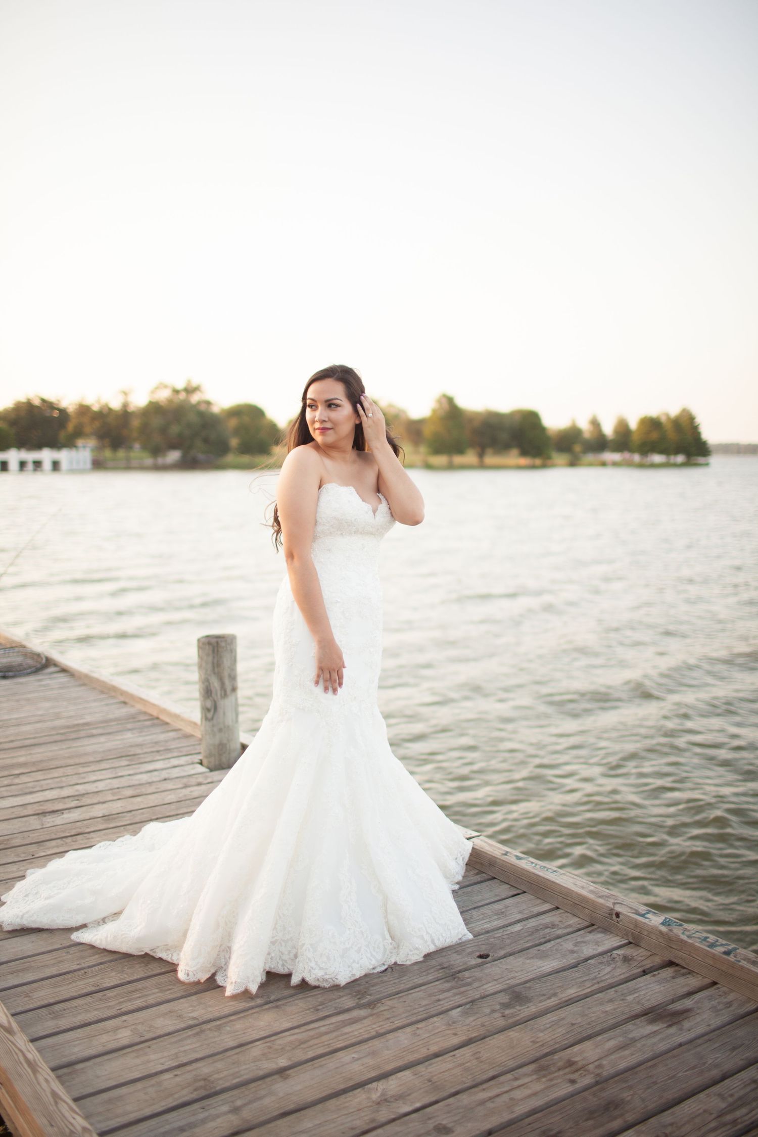 Bride in strapless lace mermaid wedding gown posing on wooden dock by lake at sunset.