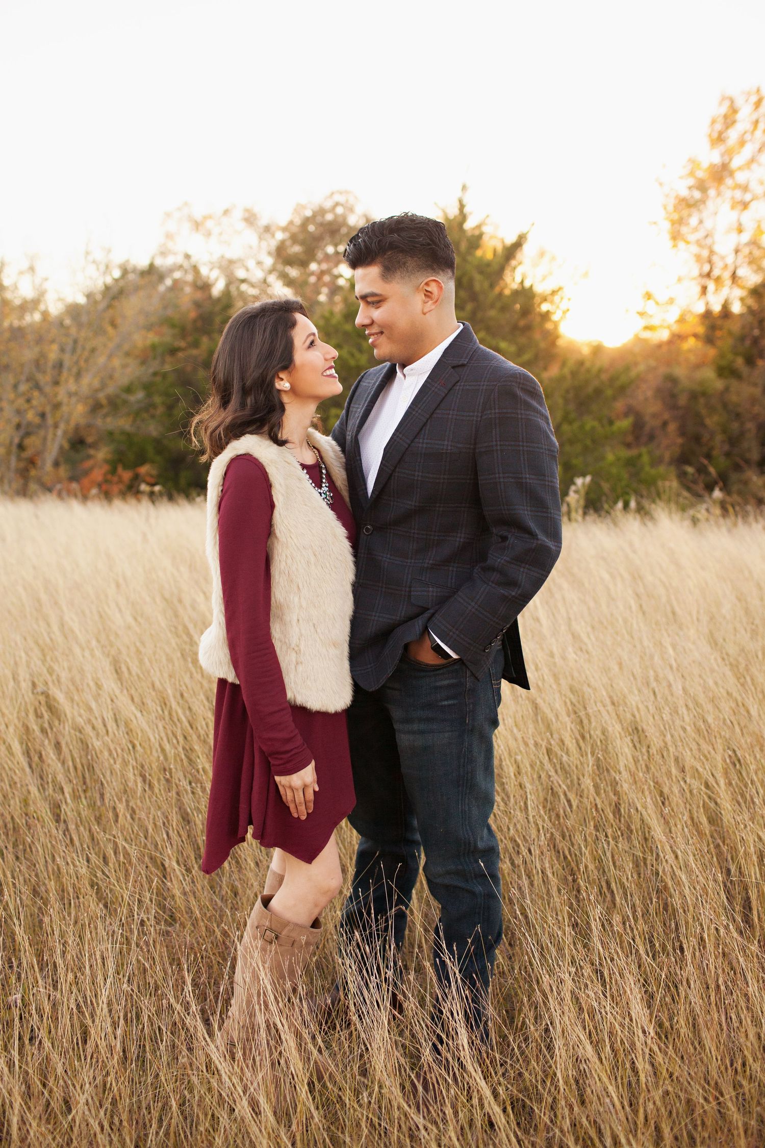 Couple sharing a romantic moment in a golden field at sunset, woman in burgundy dress and fur vest, man in blazer.