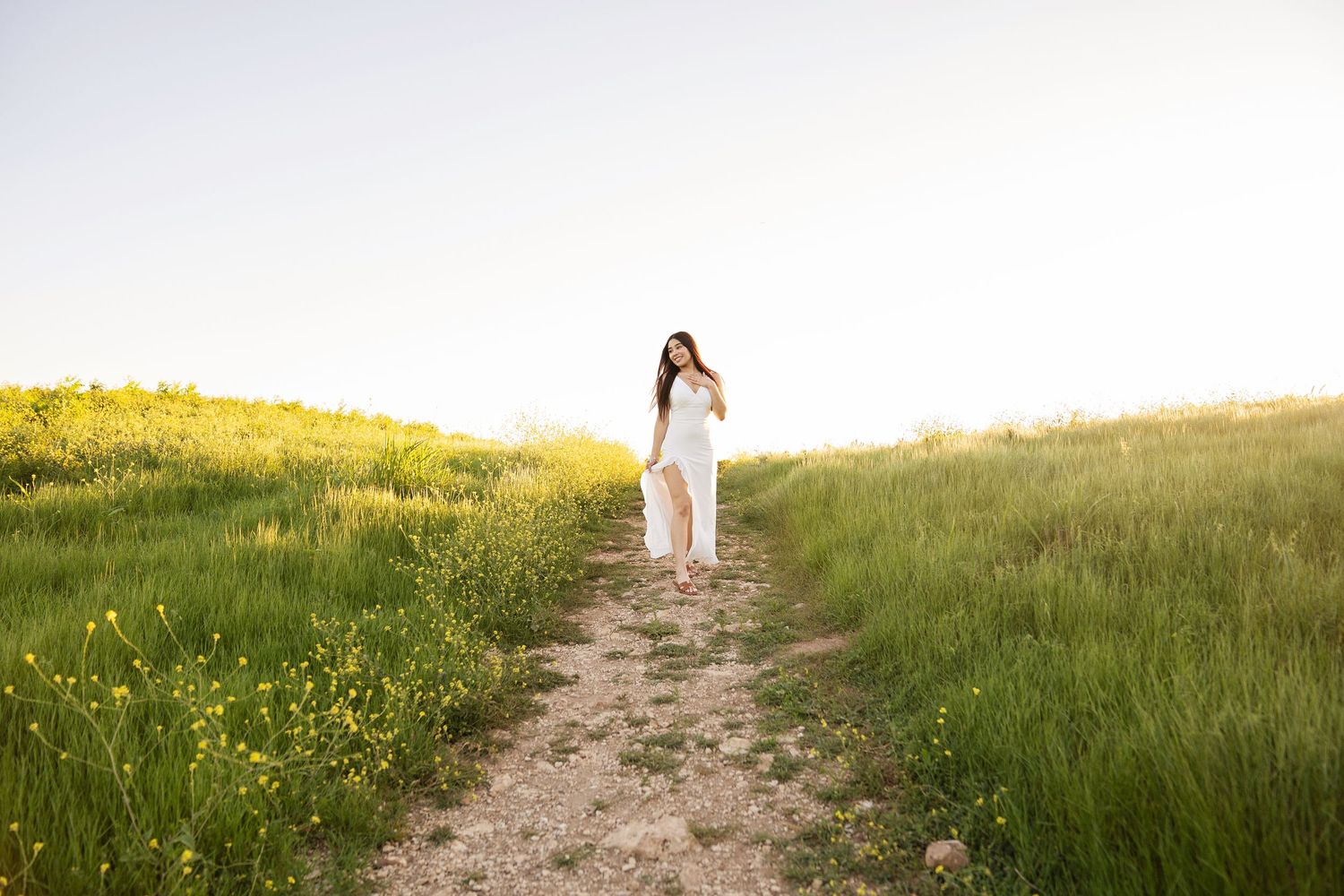 Woman in white dress walking on a dirt path through green meadow with yellow wildflowers at sunset.