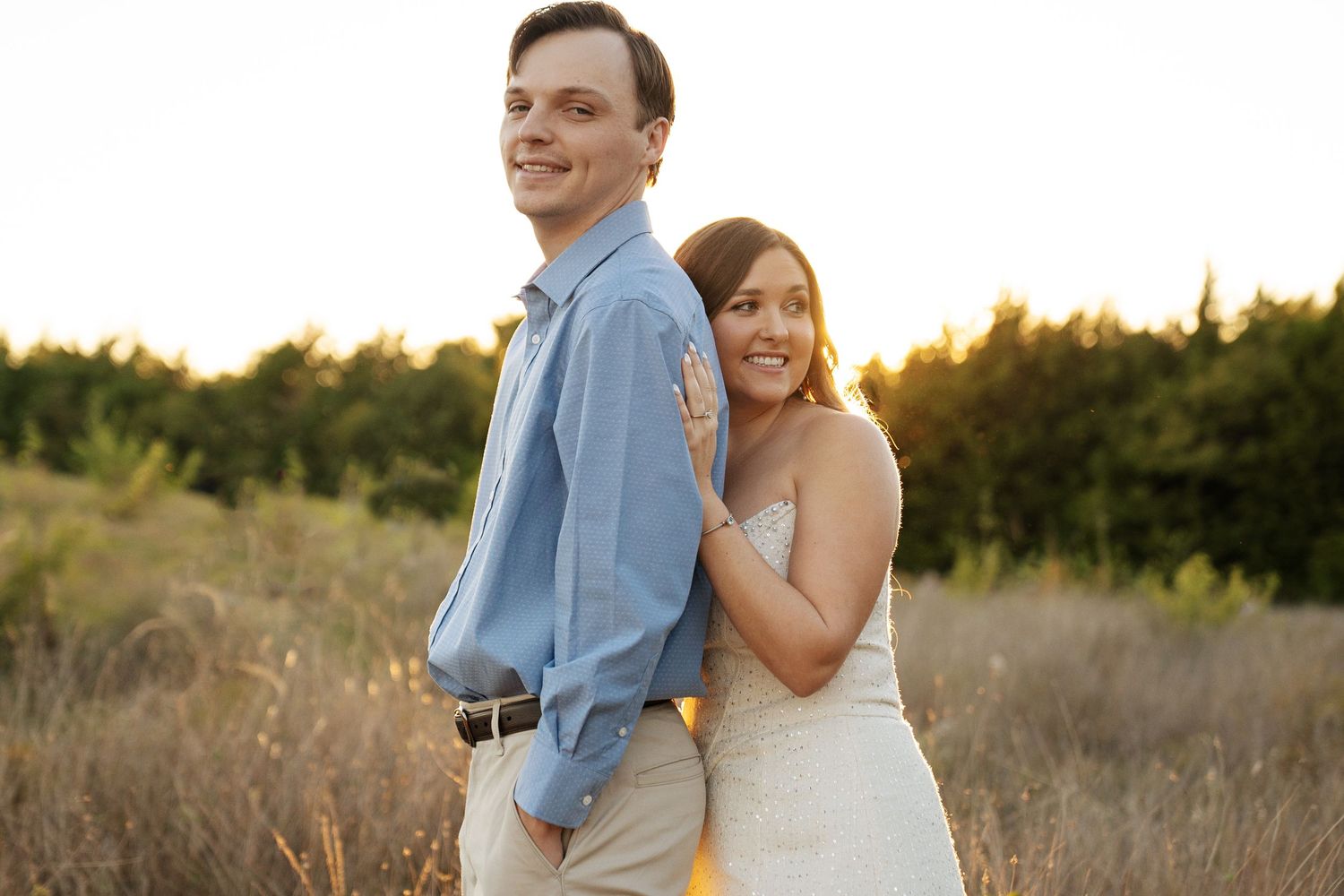Couple posing at golden hour in a field, bride in white dress hugging groom in blue shirt, trees in background.