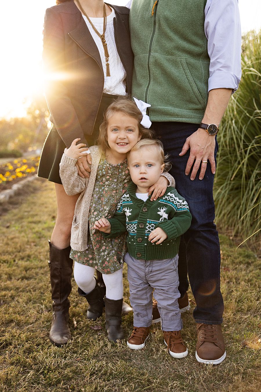 Family of four posing outdoors at golden hour, two young children in festive green sweaters with parents in a sunlit field.