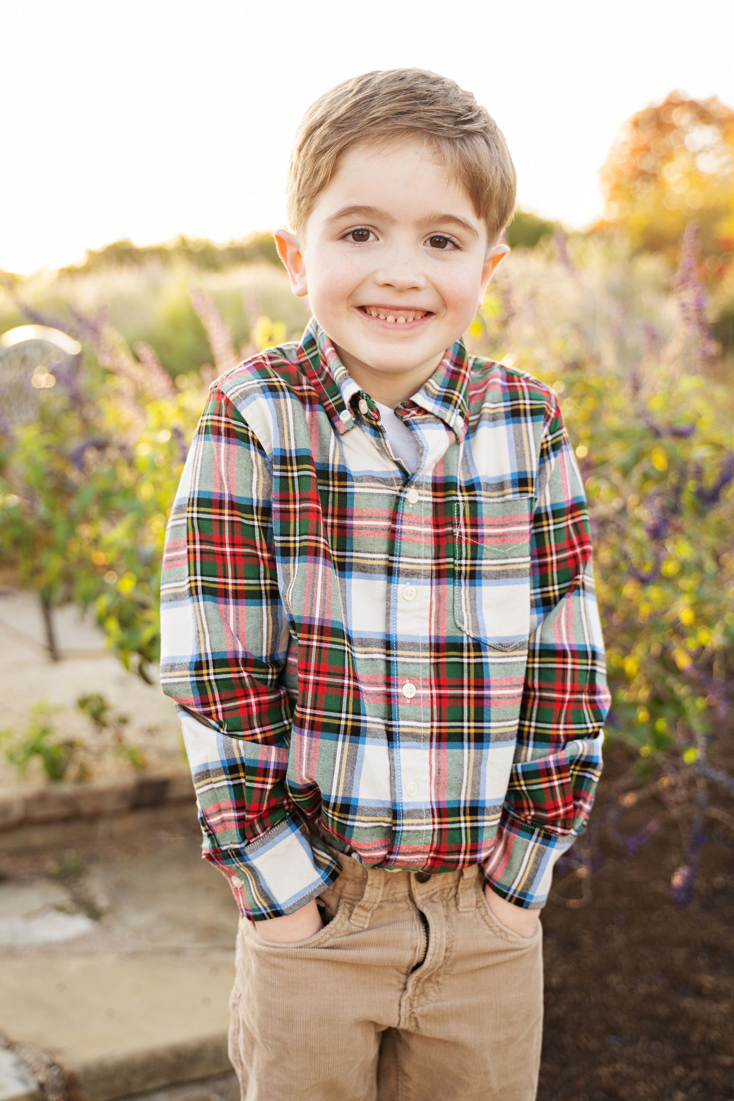 Smiling young boy in plaid shirt and khaki pants standing outdoors in a garden with colorful flowers.