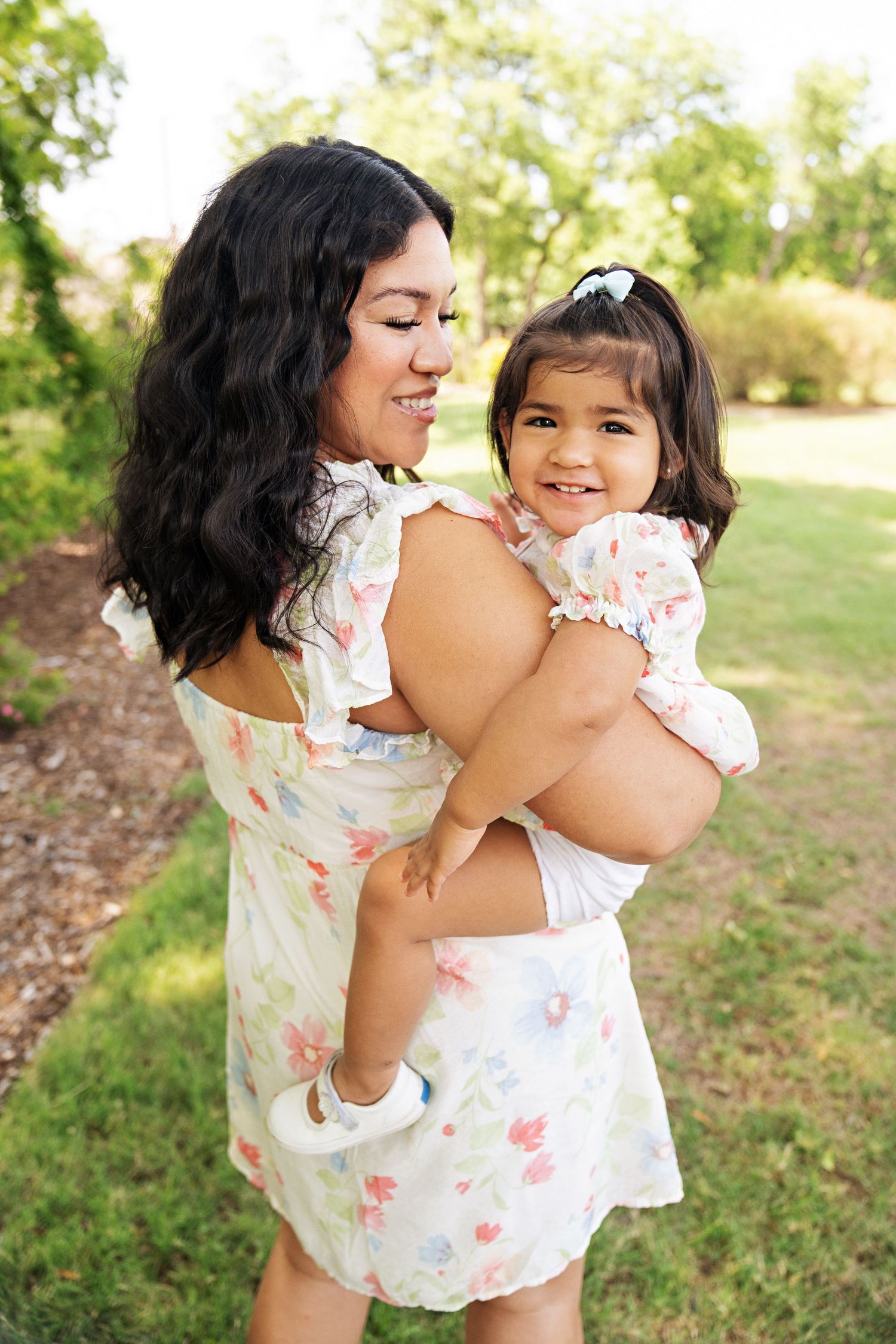 A mother in a floral dress holds her smiling toddler daughter outdoors in a lush green park setting.