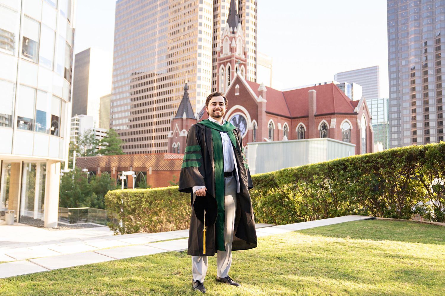 Graduate in black and green academic regalia stands on lawn with city skyscrapers and historic red church behind.