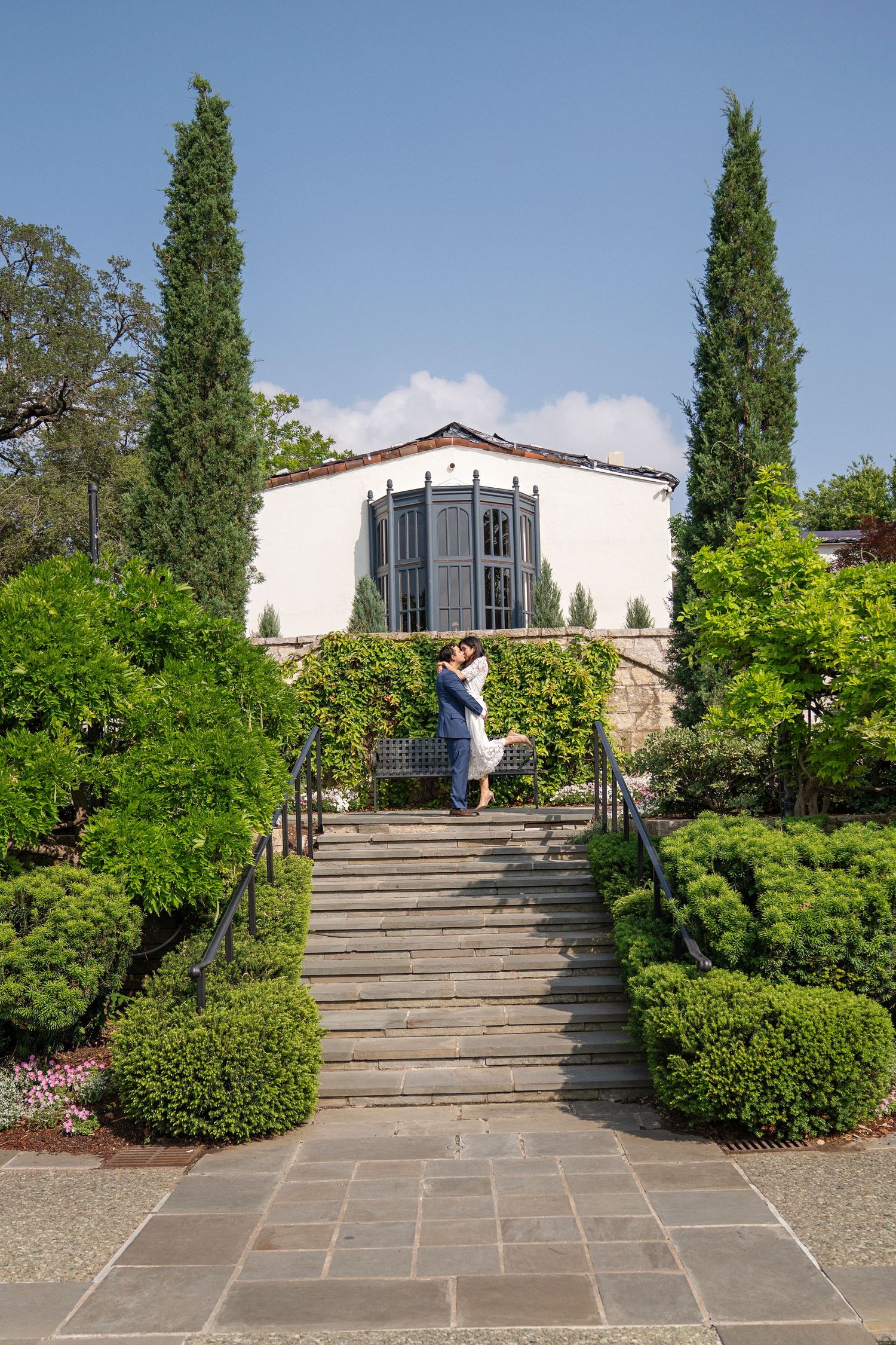 Couple embracing on stone steps in a lush garden with cypress trees and white Mediterranean-style building backdrop.