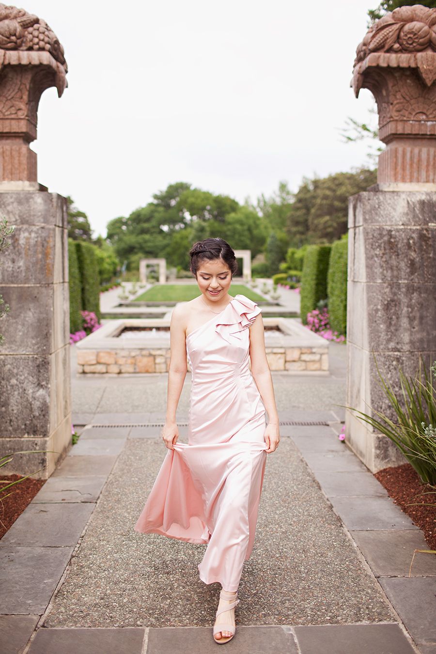 Woman in elegant pink one-shoulder satin gown walking through formal garden pathway flanked by ornate stone pillars.